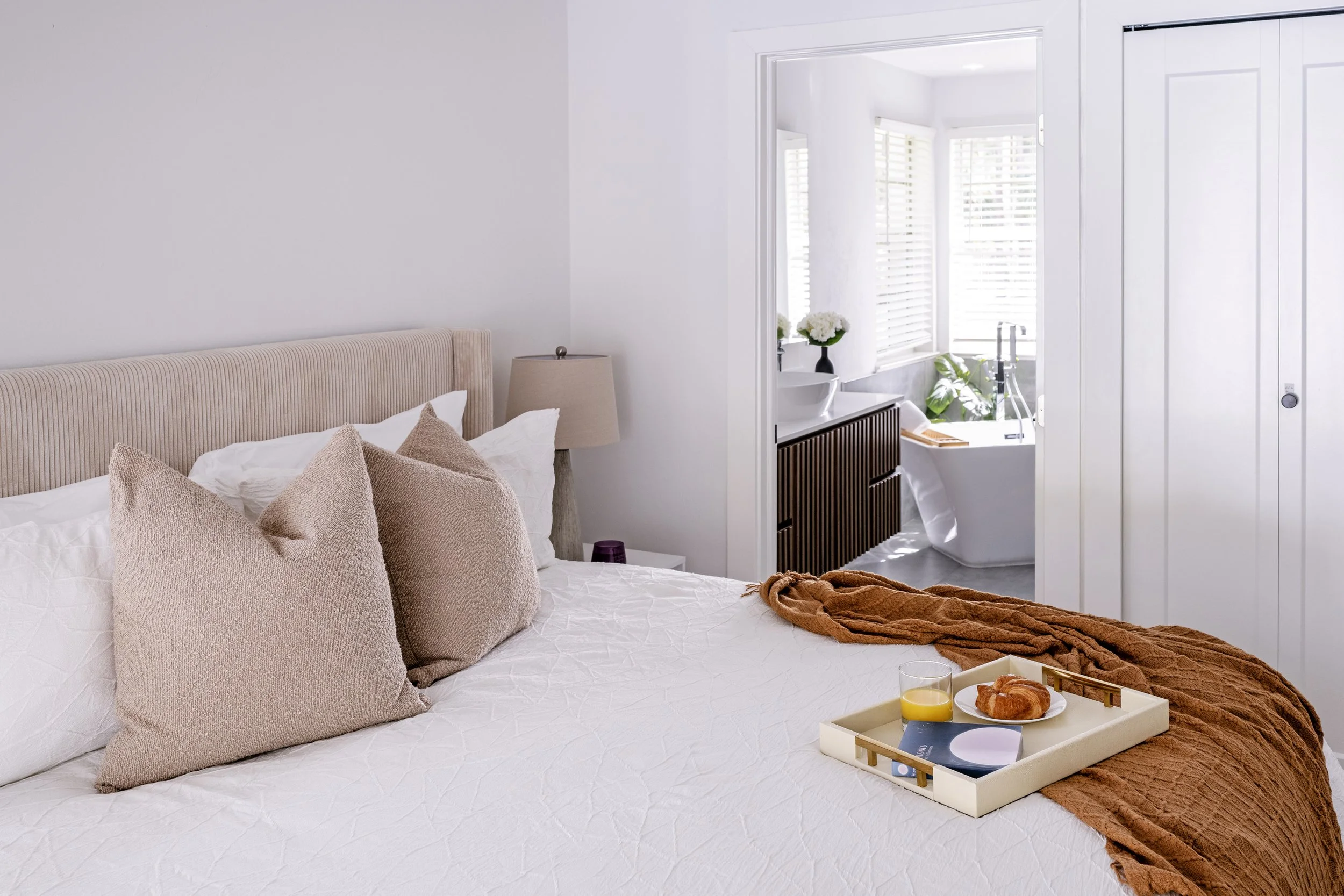 A neatly made bed with beige pillows and a brown throw blanket, a tray with breakfast items including croissants and orange juice, in a bright bedroom with white walls and a bathroom visible through an open door.