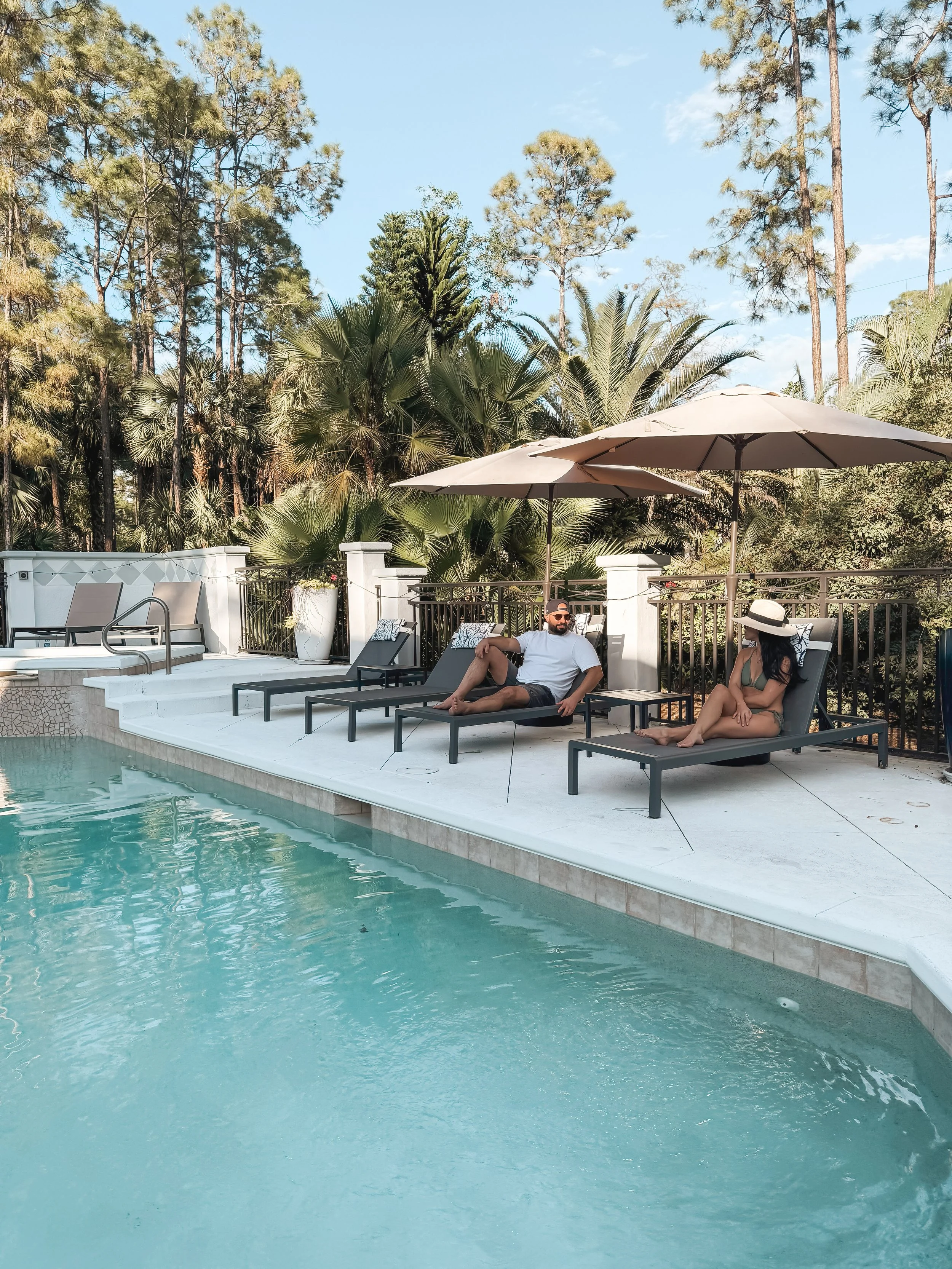 A swimming pool area with two lounge chairs with cushions, umbrellas, and a man and woman relaxing, surrounded by lush green trees and a bright blue sky.