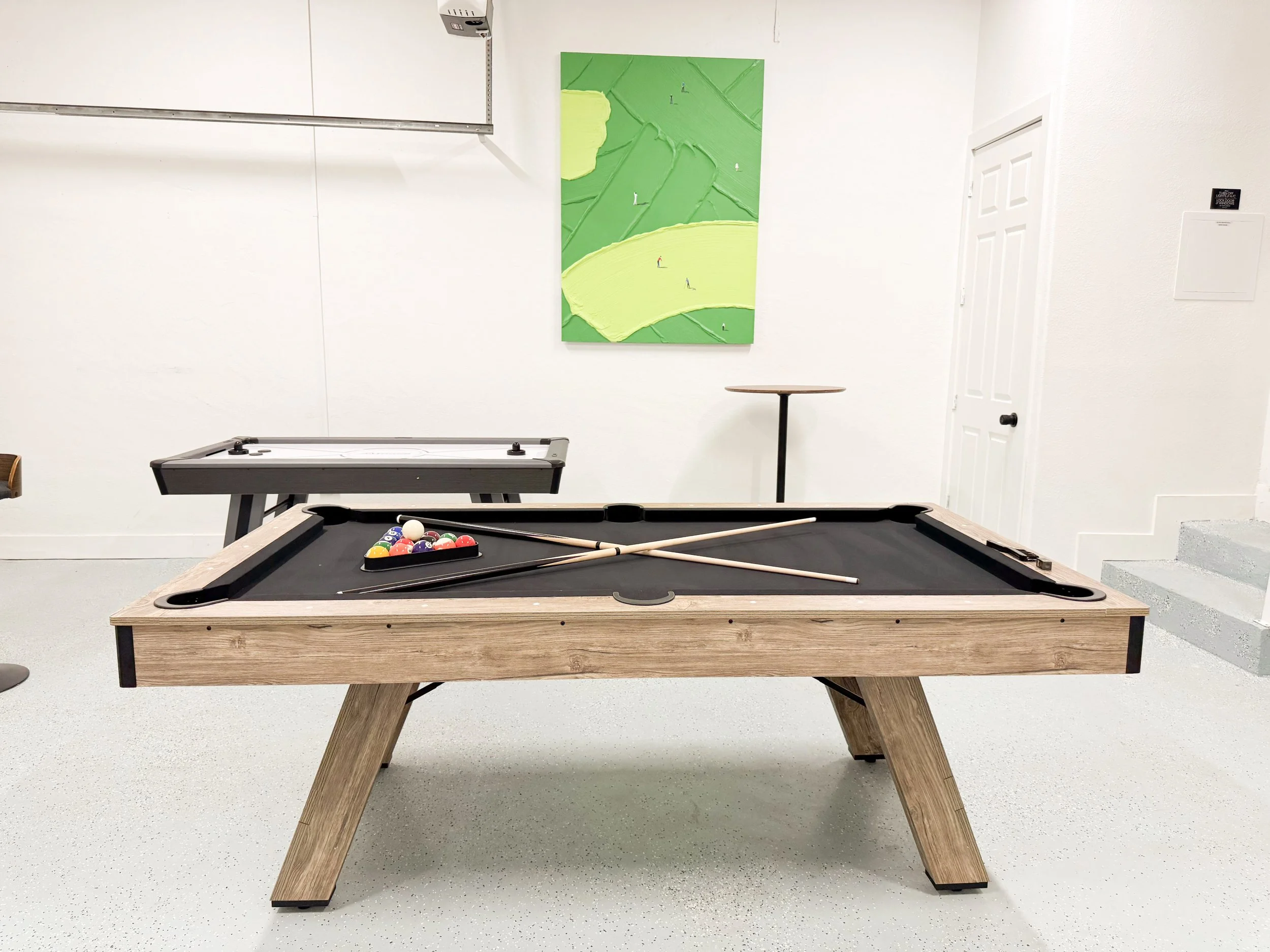 A billiards table with several pool balls arranged in a triangle, cue sticks placed on the table, in a room with white walls, a green abstract painting, and an air conditioning unit.