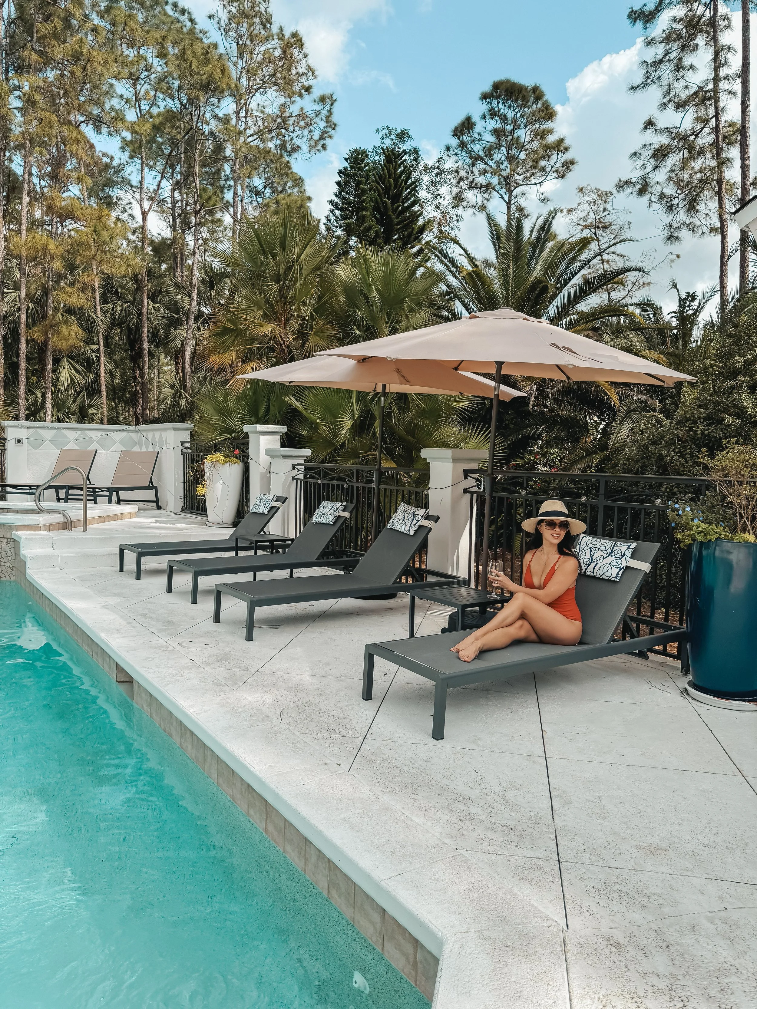 A woman in a red swimsuit and large sun hat sitting on a lounge chair by a swimming pool, holding a drink, with umbrellas and lush palm trees in the background.