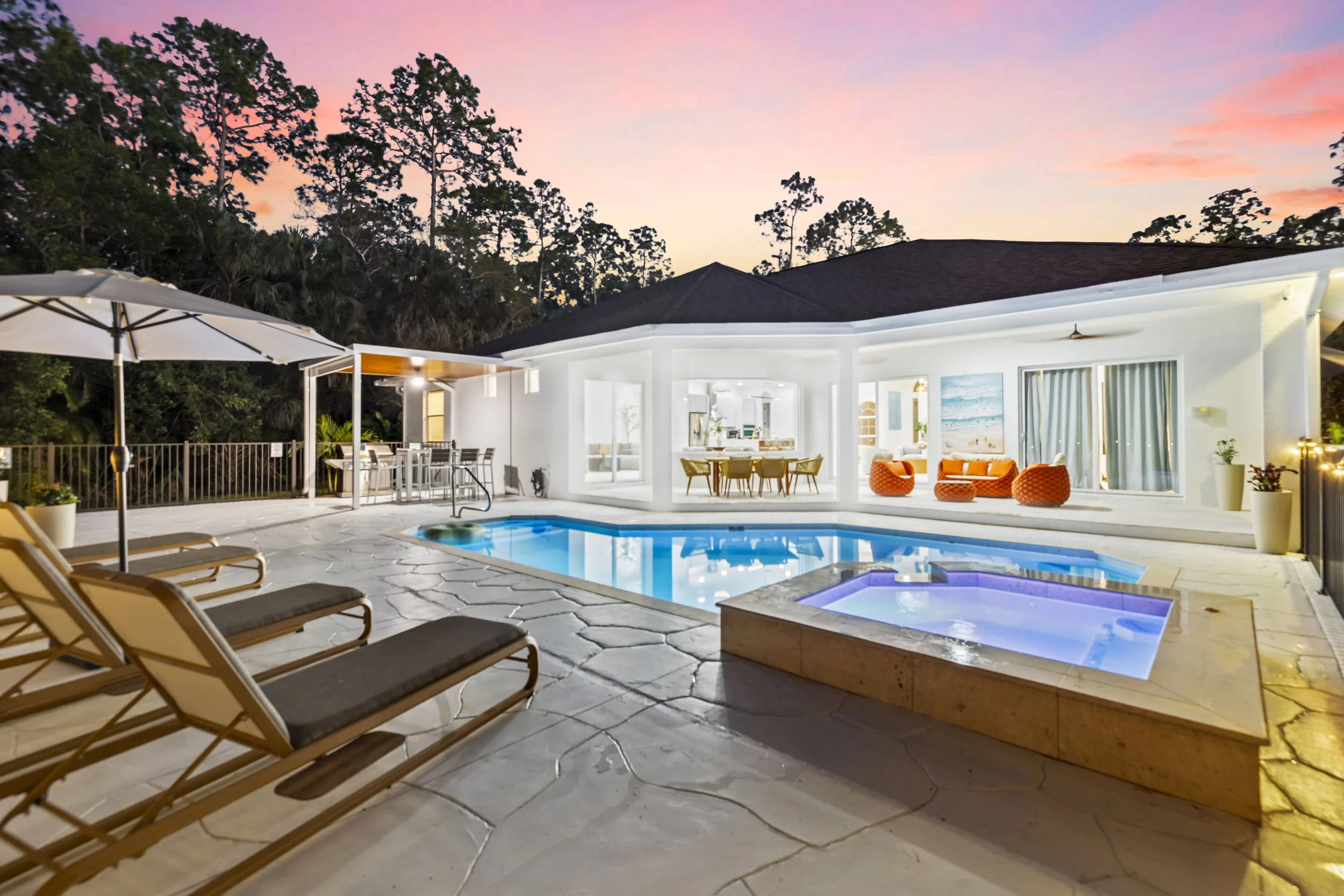 A backyard with a swimming pool and hot tub, lounge chairs, patio dining area, and outdoor seating under a covered patio at sunset, surrounded by trees.