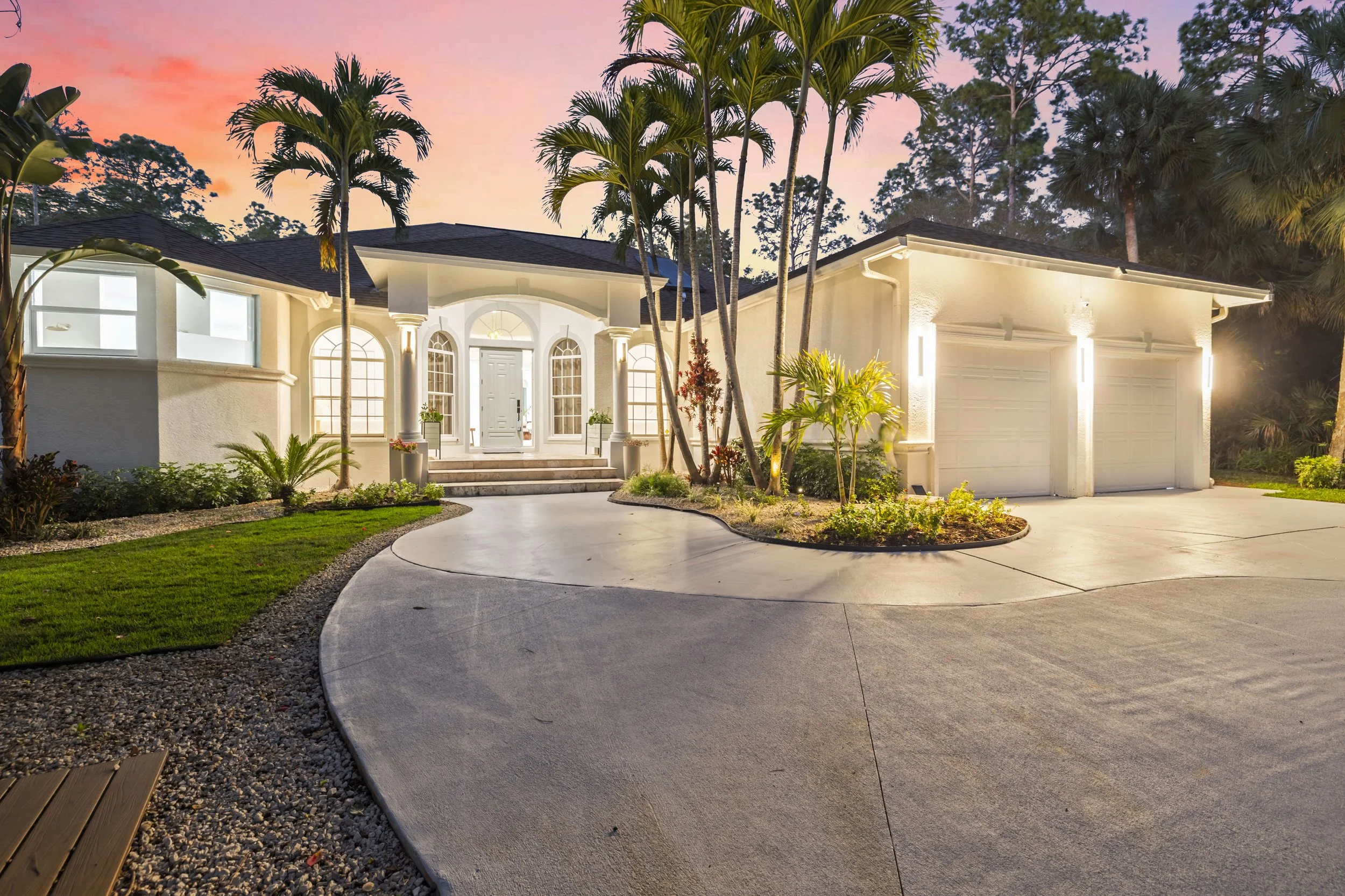 A well-lit two-story house with a white exterior, large arched windows, and a front door illuminated by outdoor lighting. The house has an attached garage, and the driveway curves around a landscaped yard with green grass, palm trees, and small plant