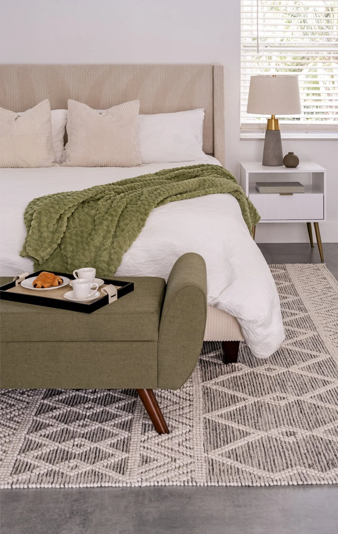 A neatly made bed with beige headboard, white bedding, beige pillows, and a green textured blanket. Next to the bed is a white nightstand with a gray and gold table lamp and small decorative items. A window with white blinds lets in light. A patterne