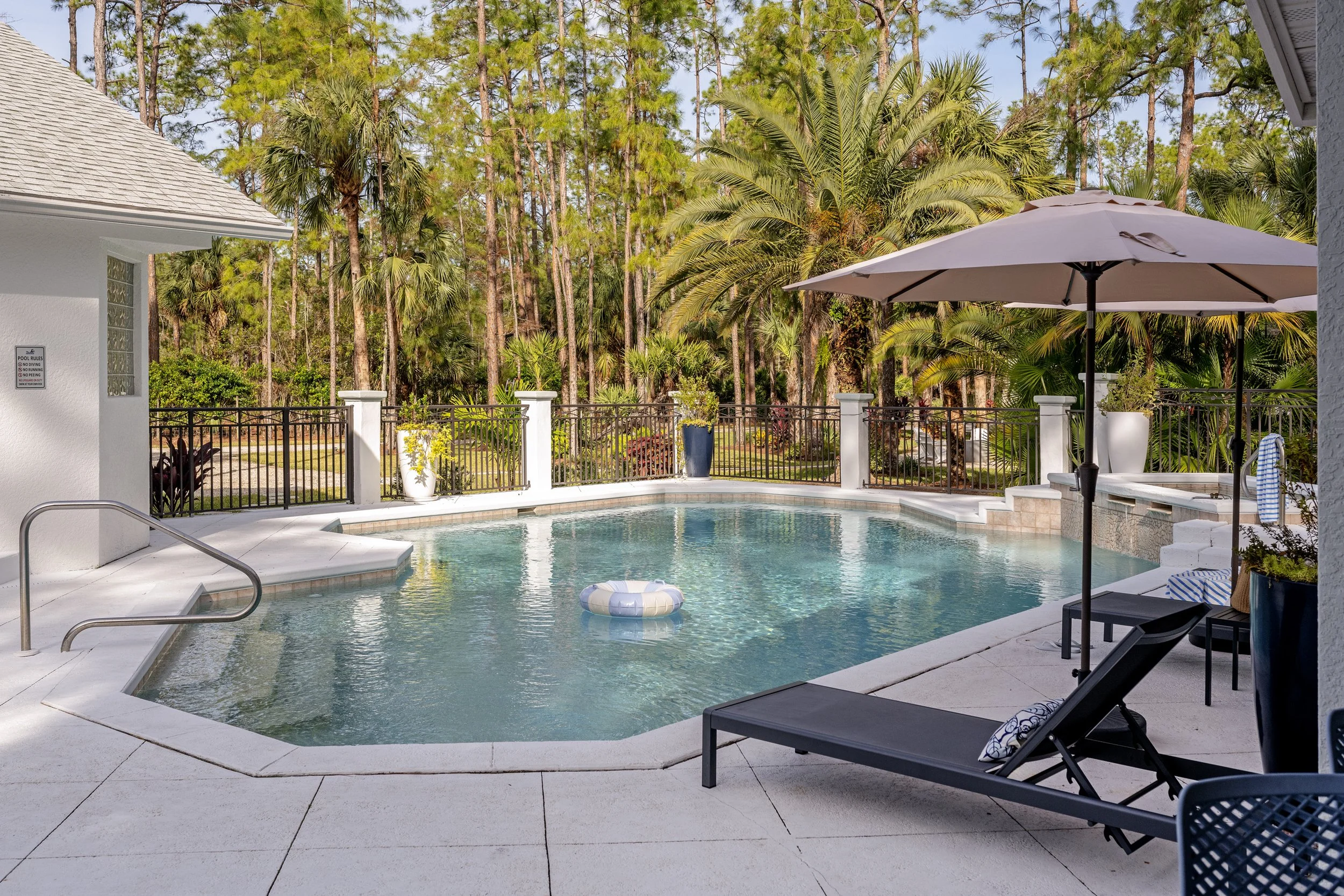 A backyard swimming pool area with lounge chairs, a large umbrella, and surrounding lush greenery including tall trees and palm plants.