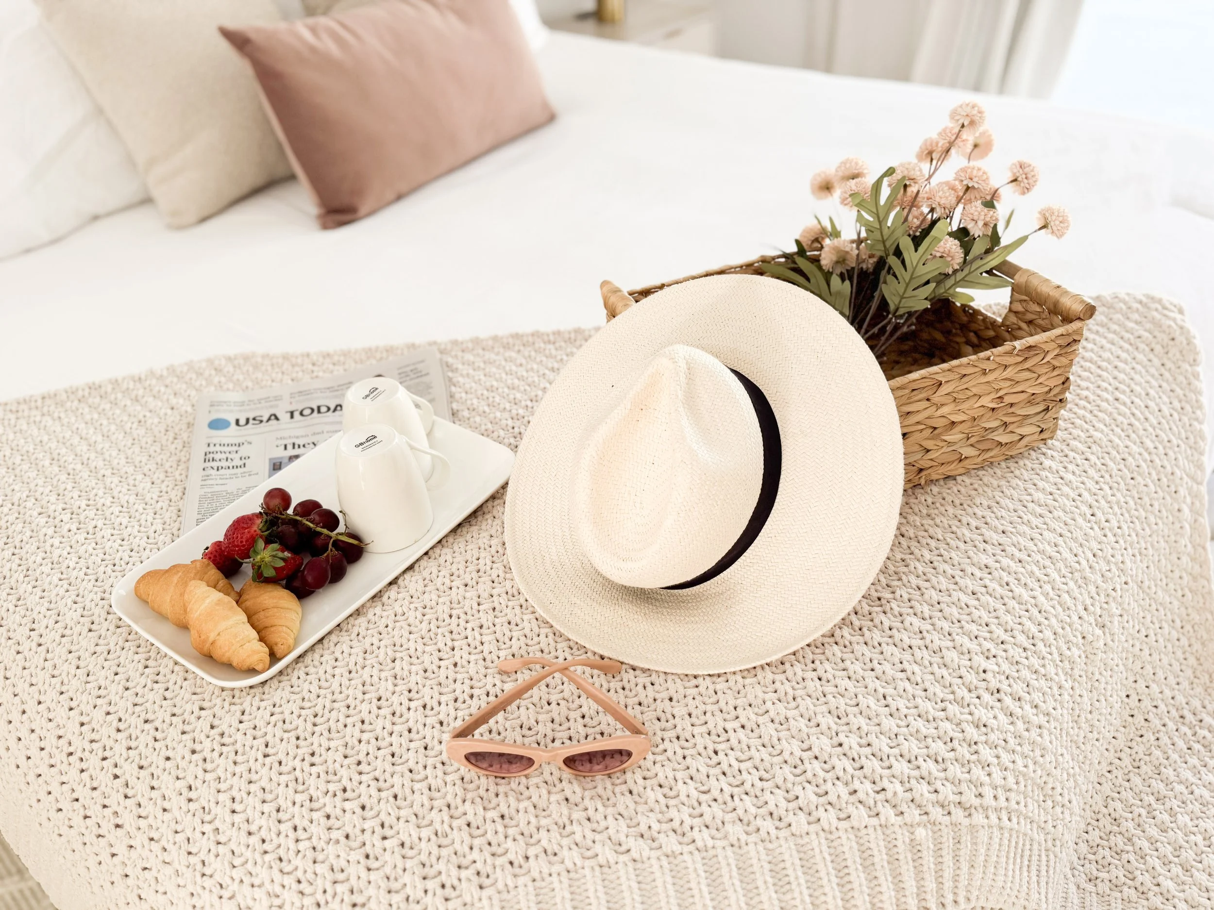 A bed with pink and cream pillows, a strip of a newspaper or magazine, a tray of croissants, strawberries, grapes, two small milk jugs, a beige hat with a black band, pink sunglasses with rose-colored lenses, and a woven basket of pink flowers on a t
