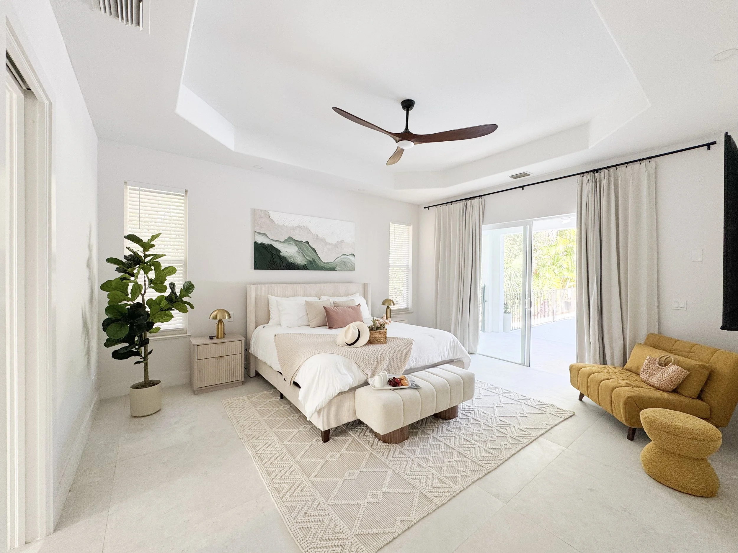 A modern bedroom with white walls and ceiling, a ceiling fan, a bed with white bedding, a beige bench at the foot of the bed, a dresser, and a large window with white curtains.
