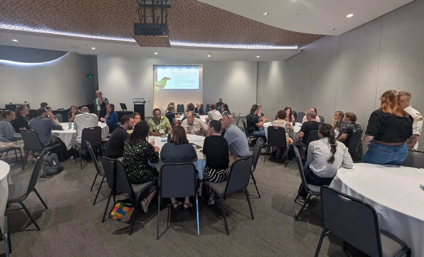 A crowded conference or seminar room with multiple round tables and people engaged in discussions. Some individuals are standing, others are seated, and a presentation is displayed on a screen at the front of the room.