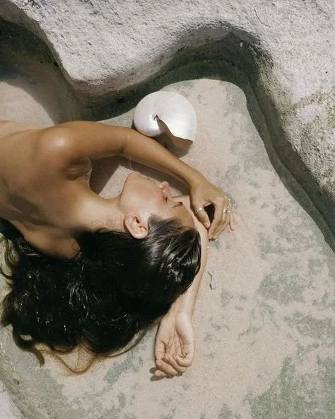 A woman with long dark hair lying on a flat surface, resting her head on her folded arms, next to a white coconut.