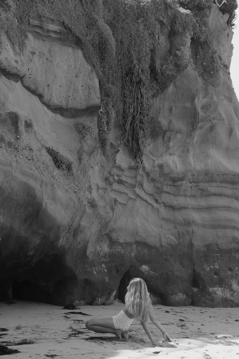 A woman with long wavy hair in a white dress crouches on a sandy beach at the base of a large rock formation with layered patterns and sparse vegetation.