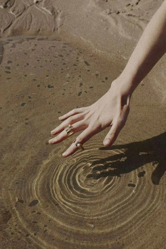 A person's hand with rings touching wet sand, creating concentric ripples near the shoreline.