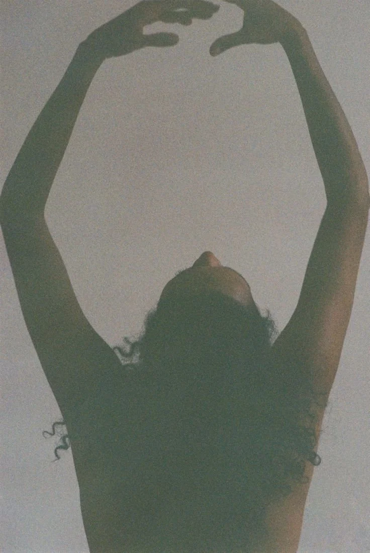 A person with curly hair was captured from below, with their arms raised above their head and fingers forming a shape against the ceiling or sky.