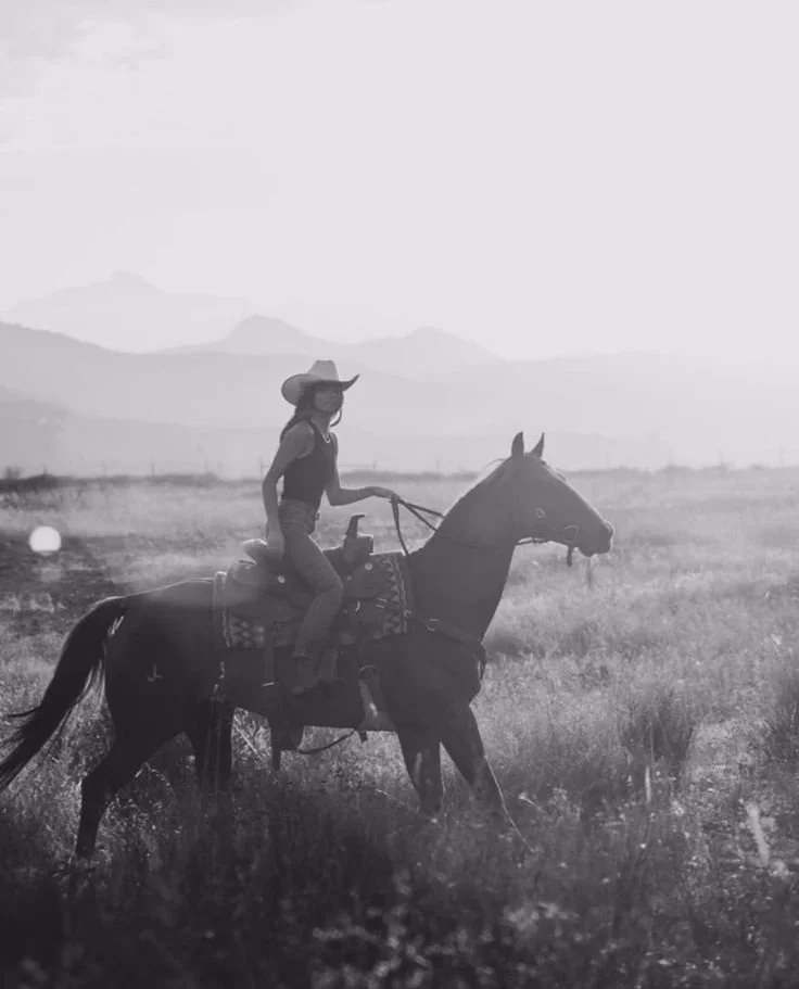 A woman wearing a cowboy hat riding a horse through a vast open field with mountains in the background, captured in black and white.