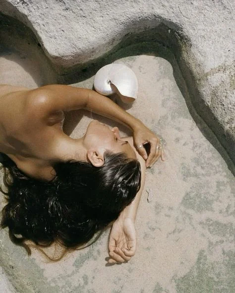 A woman with dark hair lying on her side on a rock surface, next to a broken coconut, with her arm resting on her head.