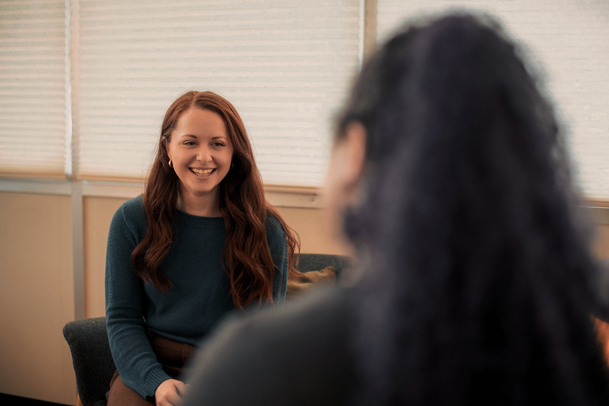 A woman with long wavy red hair smiling and wearing a teal sweater. She is sitting across from another person whose head and hair are visible in the foreground, blurred. The background shows window blinds.