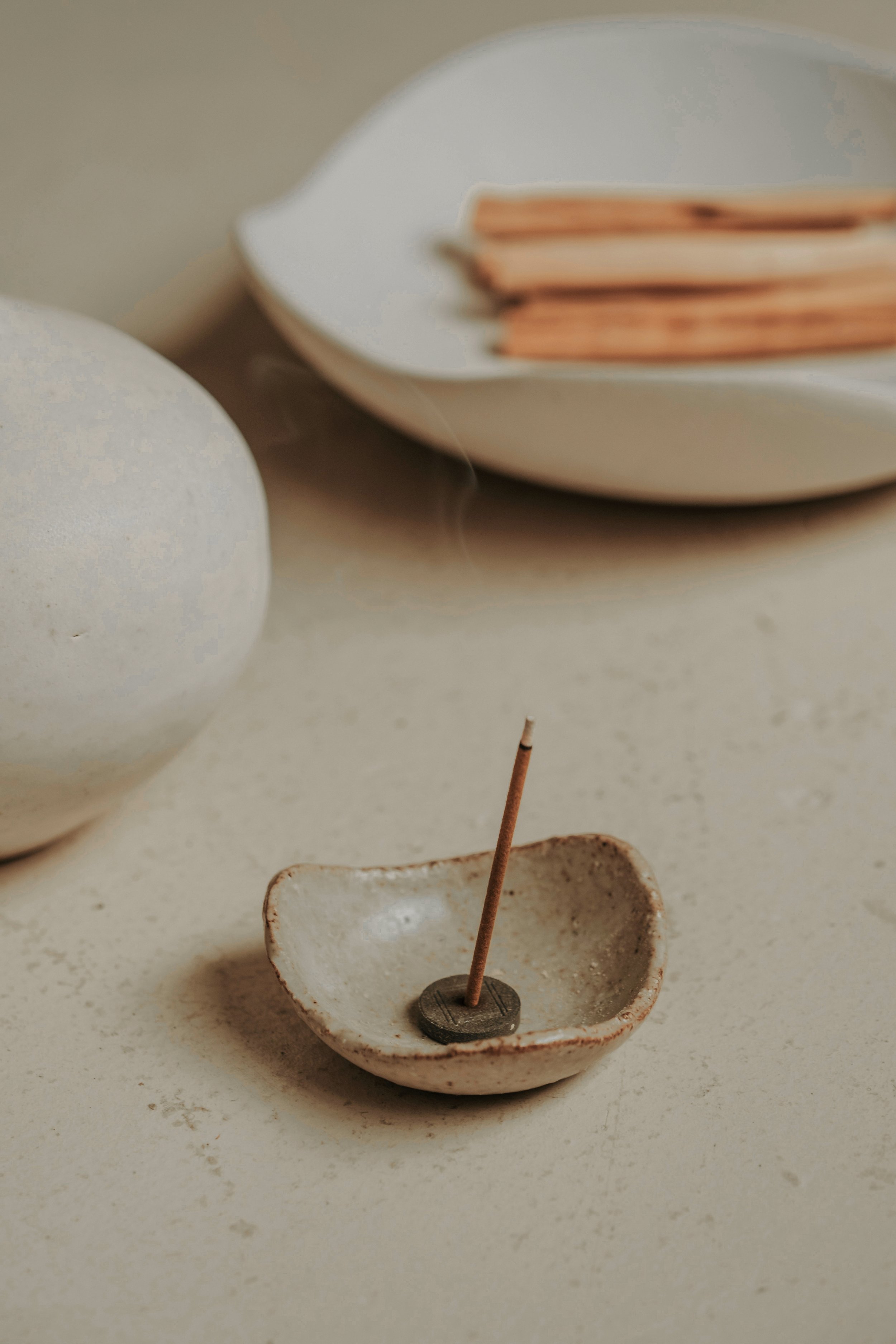 A small ceramic dish holding a stick-shaped object with a button at the base, set on a lighter-colored surface. In the background, there are two eggs and a white plate with several cinnamon sticks.