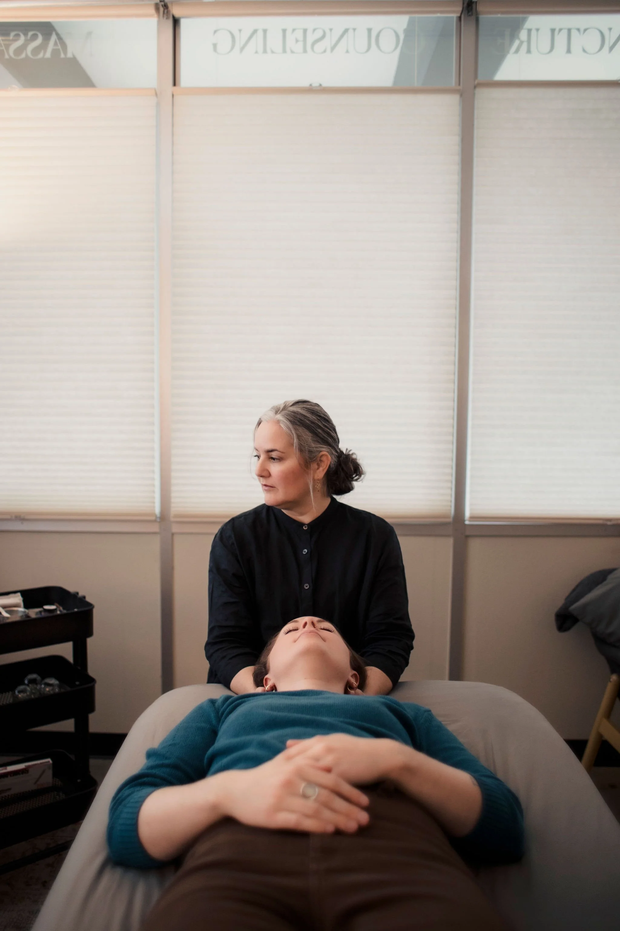 A woman receiving a facial treatment while lying on a treatment table, with a technician standing beside her in a room with closed blinds.