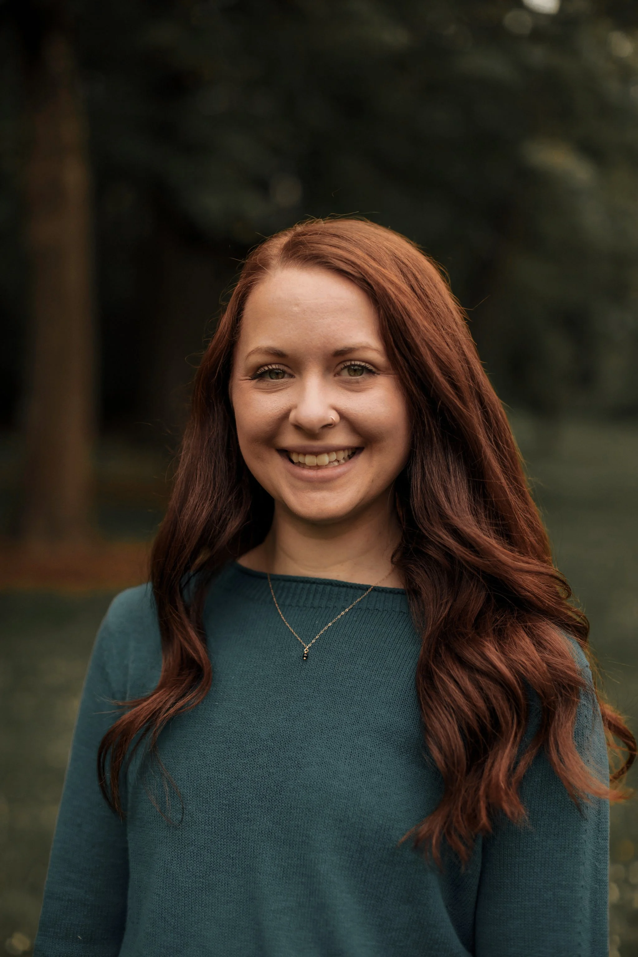 A woman with red hair smiling outdoors with a blurred forest background.