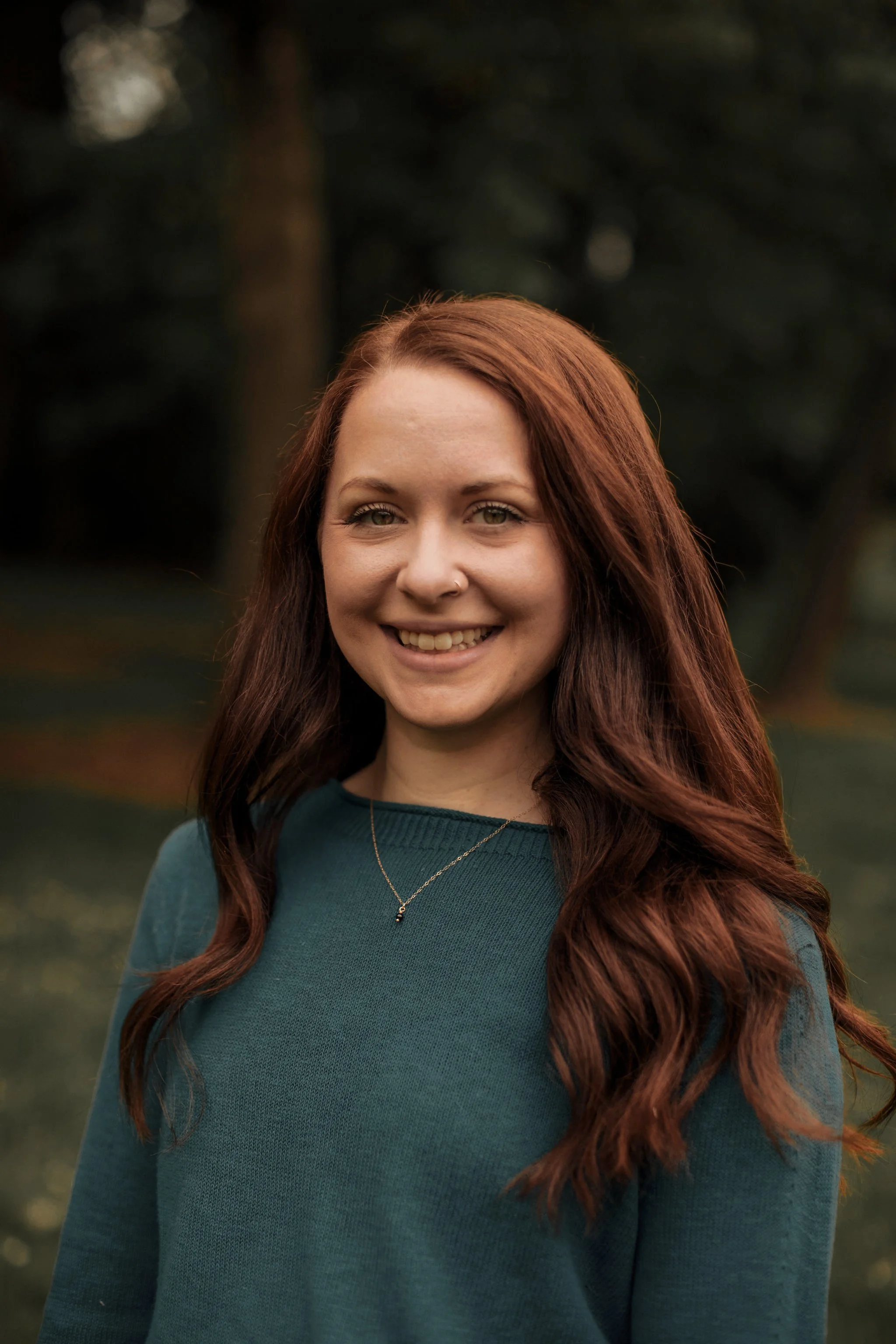 A young woman with long red hair and light skin standing outdoors in front of a dark, blurred background. She is smiling and wearing a teal long-sleeve top and a delicate necklace.