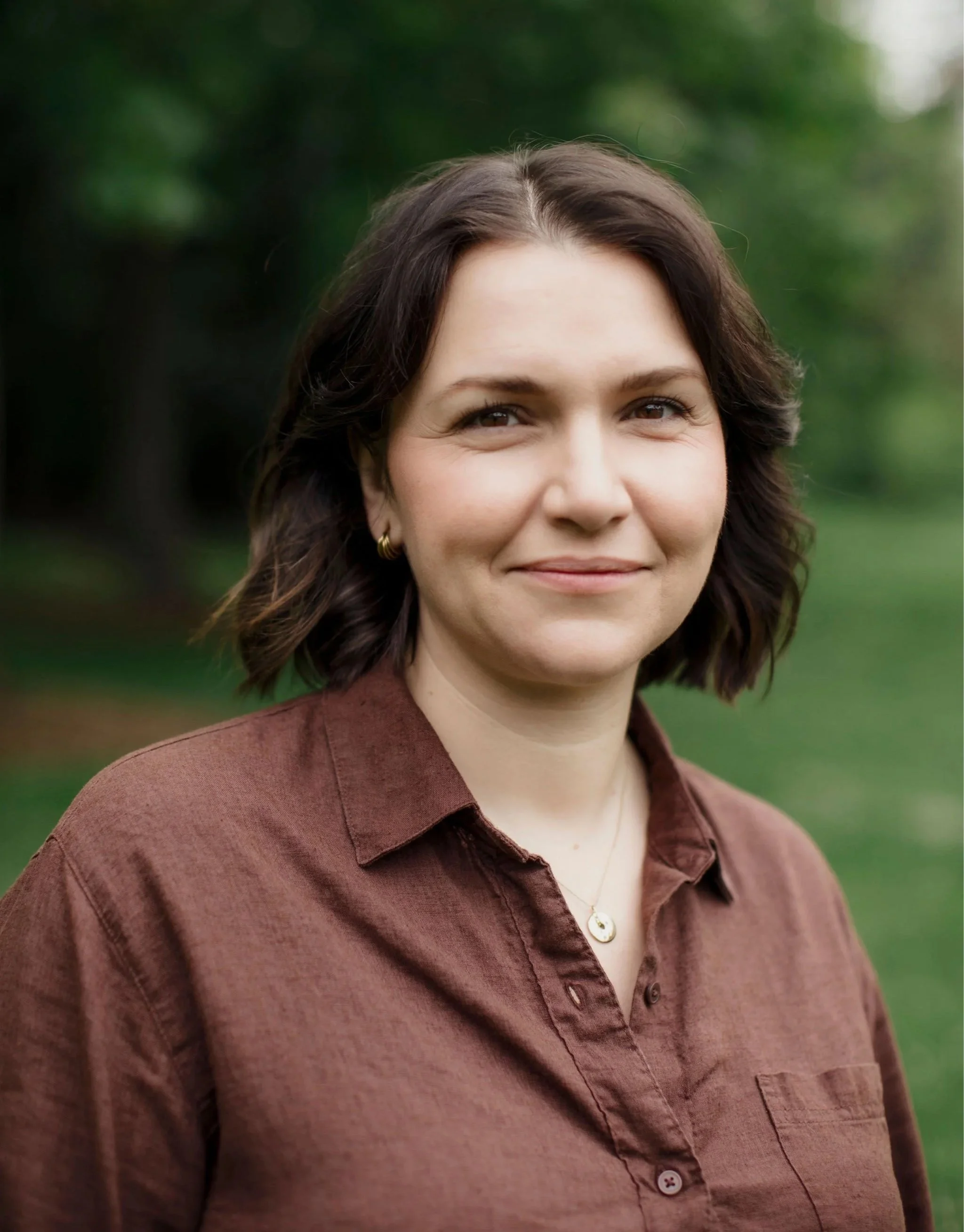 A woman with dark brown hair styled in loose waves, wearing a brown button-up shirt, standing outdoors with a blurred green background.
