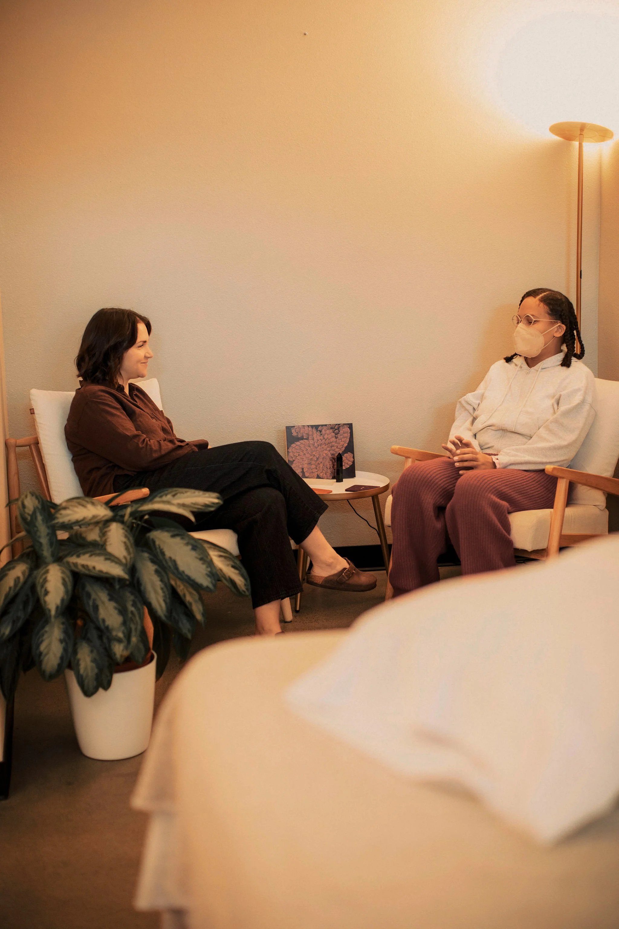 Two women sitting in a room having a conversation. One woman is wearing a brown top and black pants, the other is wearing a white hoodie and pink pants, both are wearing masks. A small table with decorative items is between them and there's a plant in the foreground.