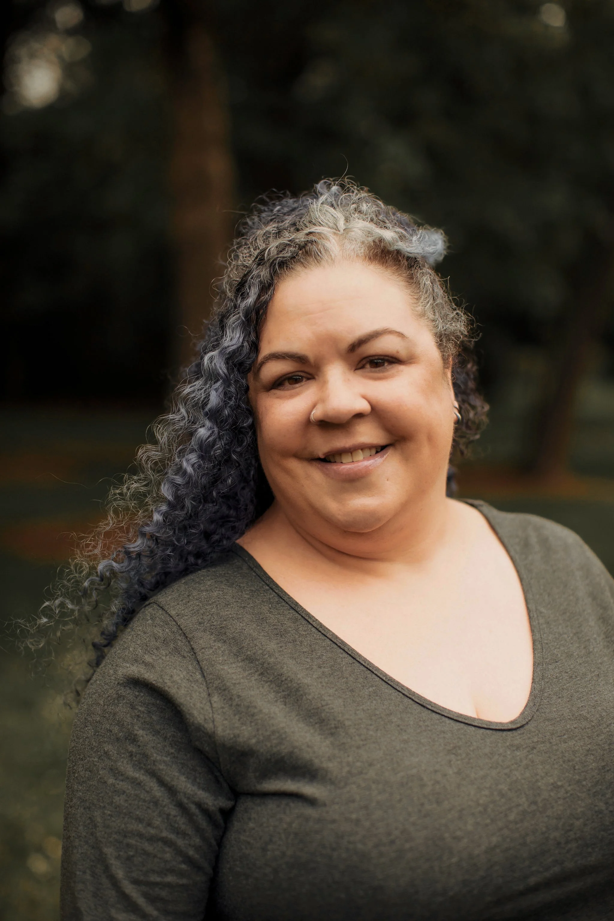 A woman with curly, salt-and-pepper hair smiling outdoors in front of blurred trees.
