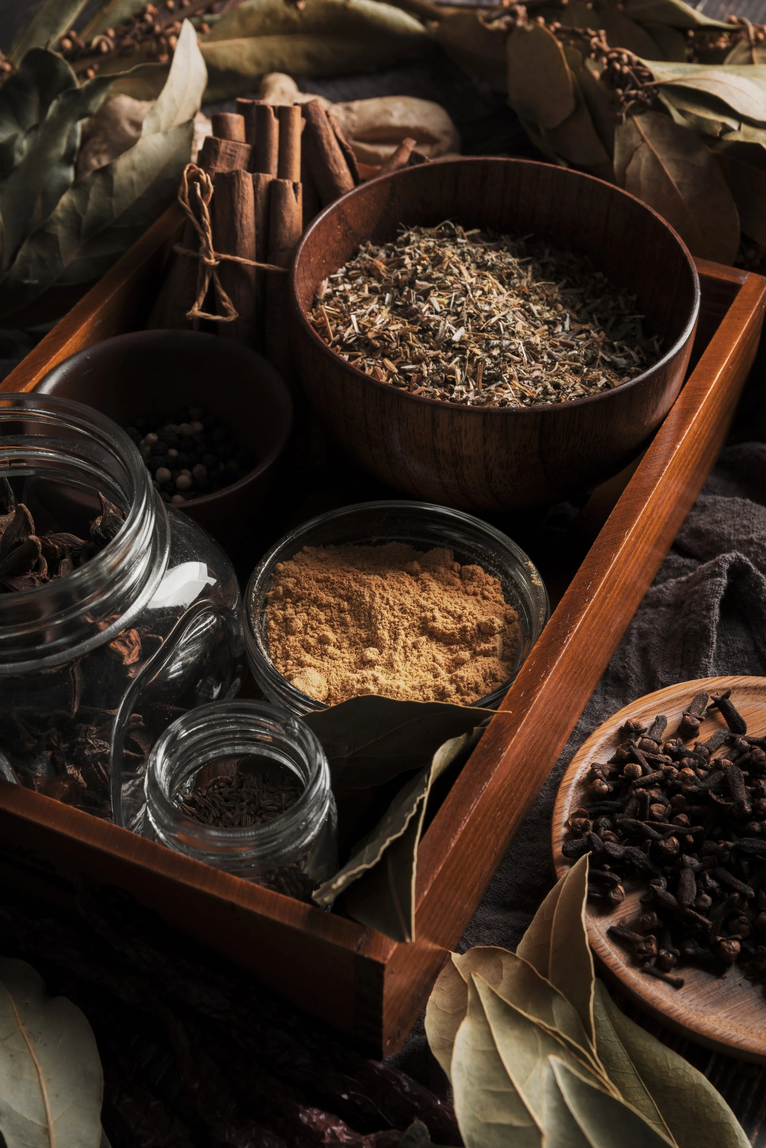 A wooden tray filled with various spices and herbs, including cinnamon sticks, dried herbs, and powder, surrounded by dried bay leaves and other small containers of spices.
