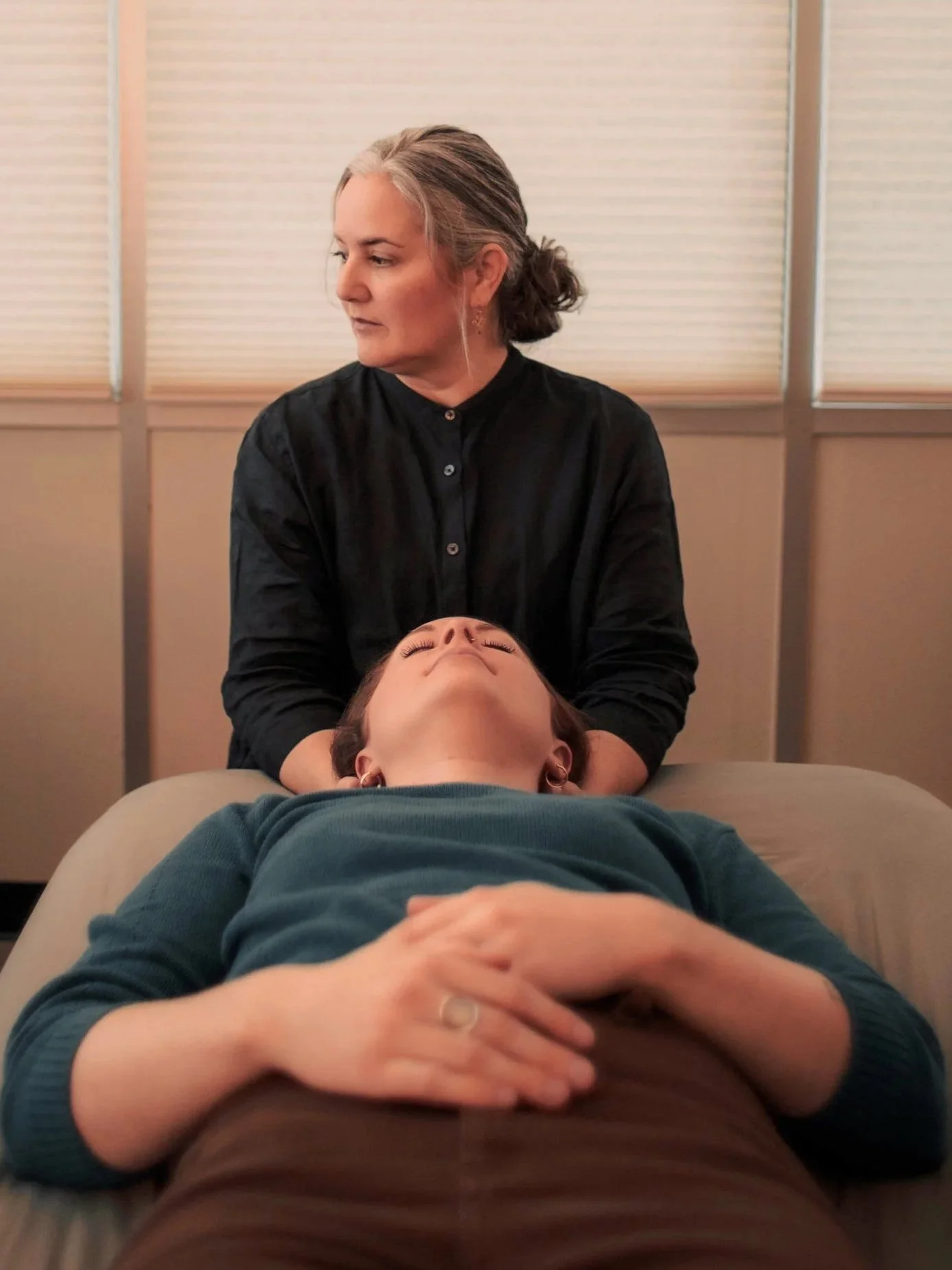 A woman with gray hair in a bun and earrings is receiving a head massage from a person lying on a massage table. The person on the table has short dark hair and is wearing a dark green top.
