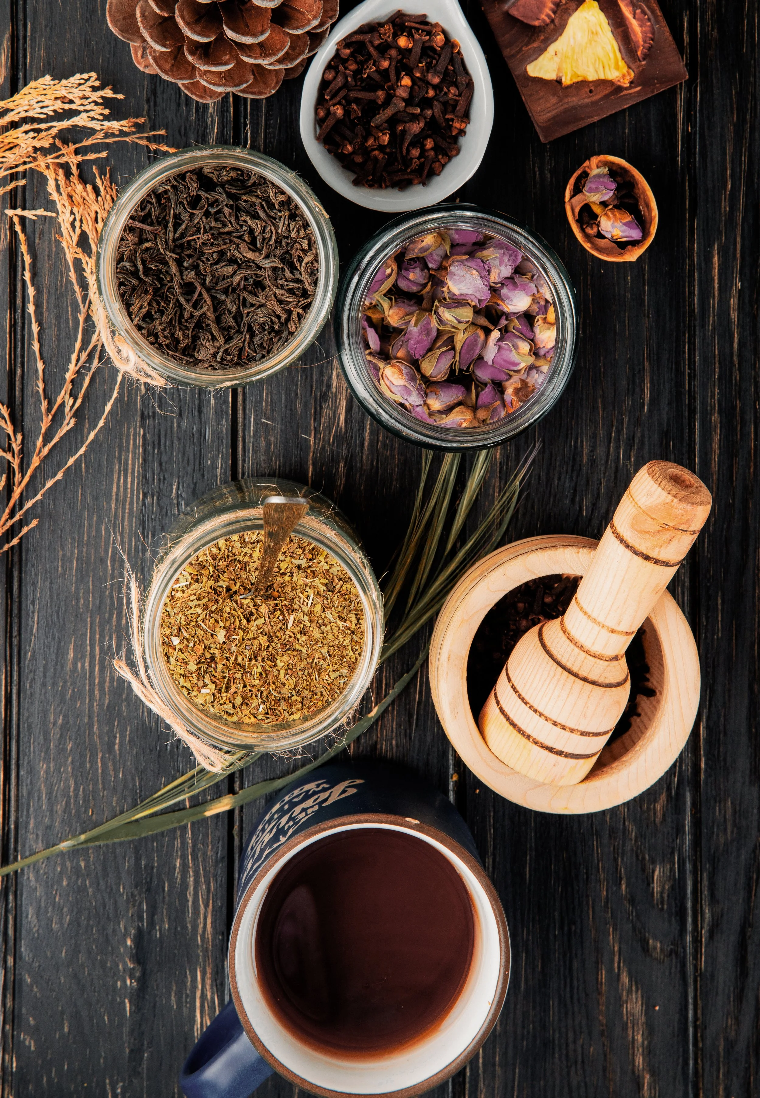Top view of a rustic wooden table with an assortment of dried herbs, tea leaves, incense, a pine cone, a cup of tea, a wooden mortar and pestle, and dried rose petals in jars.