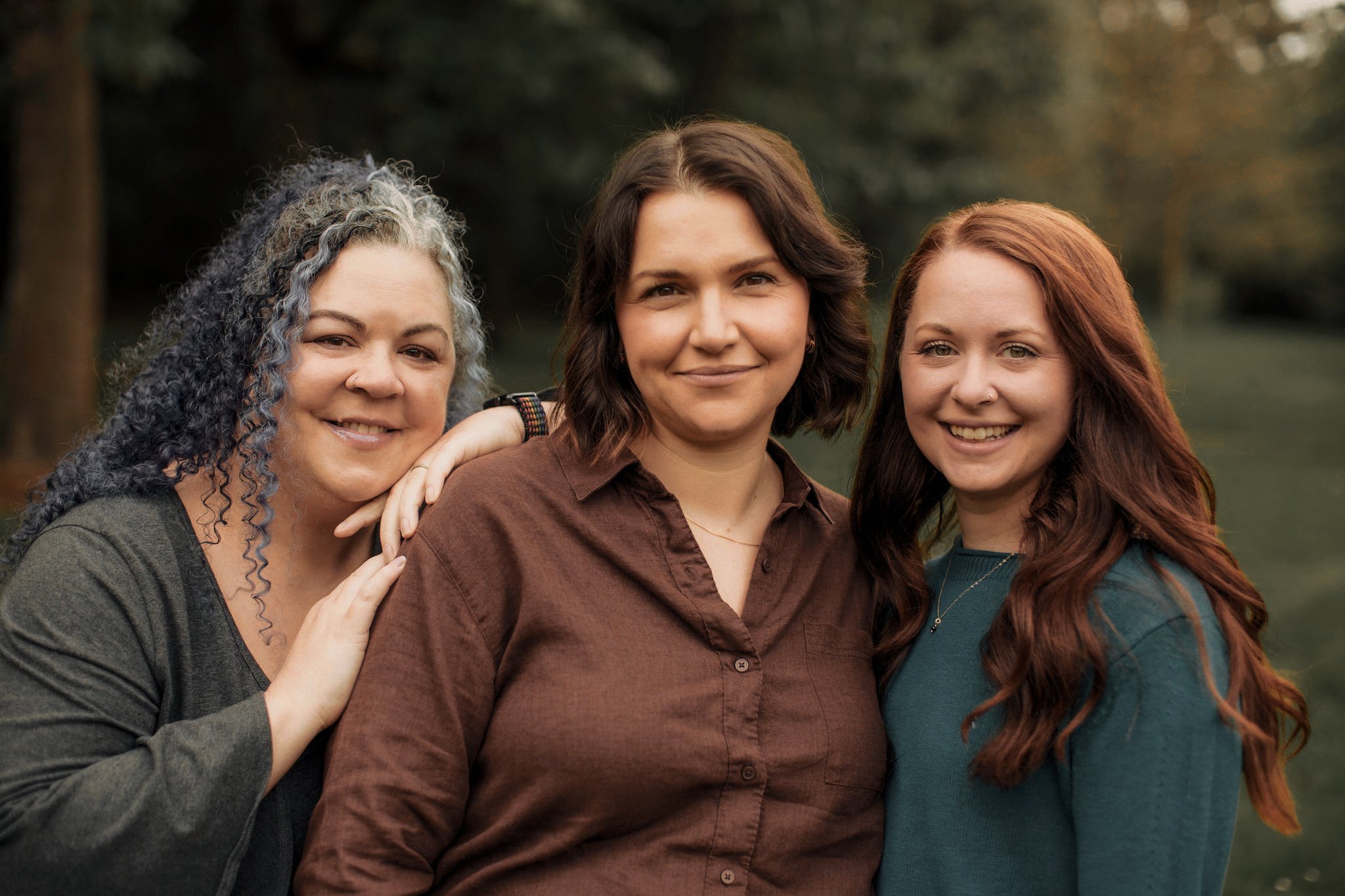 Three women smiling outdoors with trees in the background, one has gray curly hair, the second has short brown hair, and the third has long red hair.