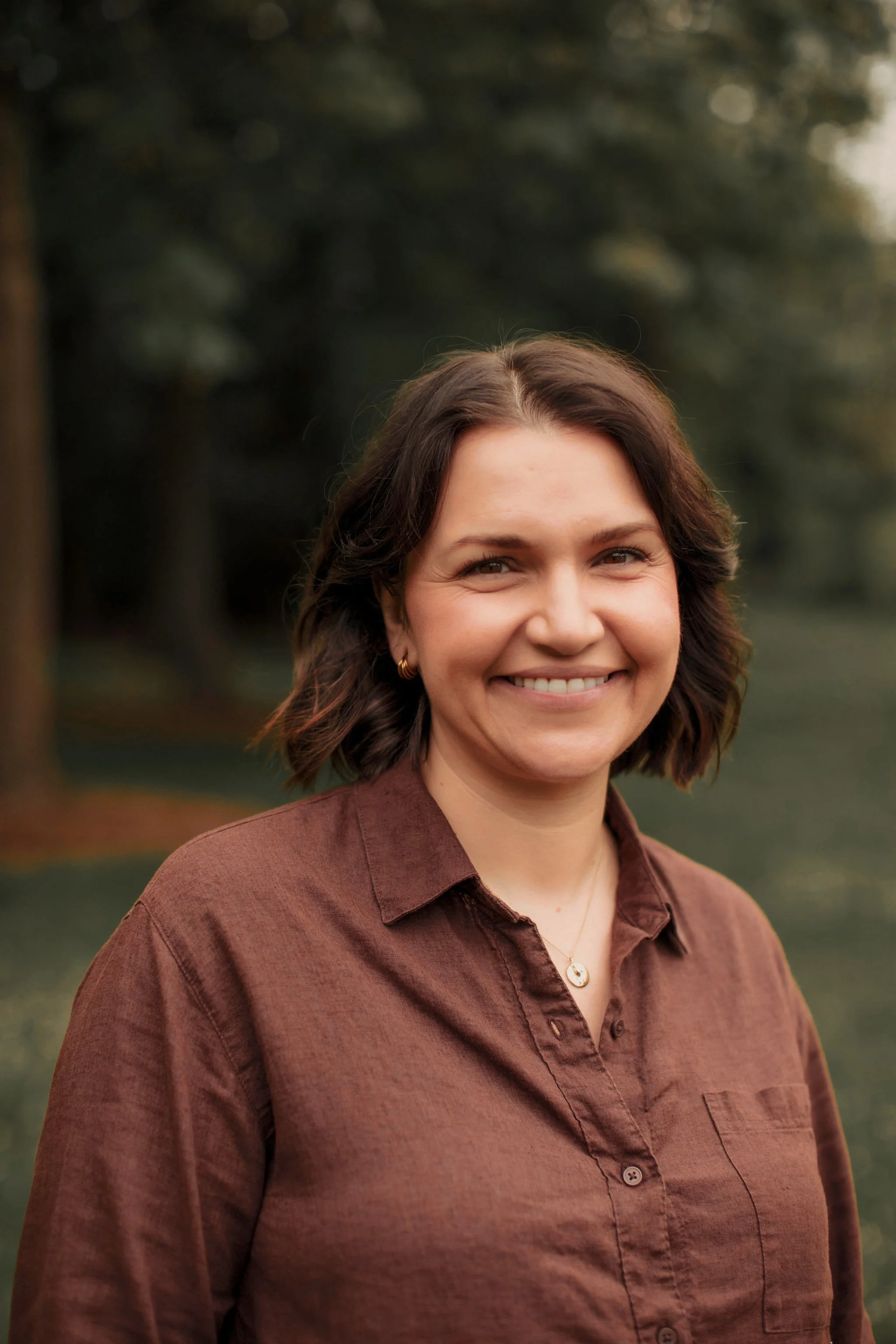 A woman with shoulder-length brown hair smiling outdoors with blurred trees in the background.