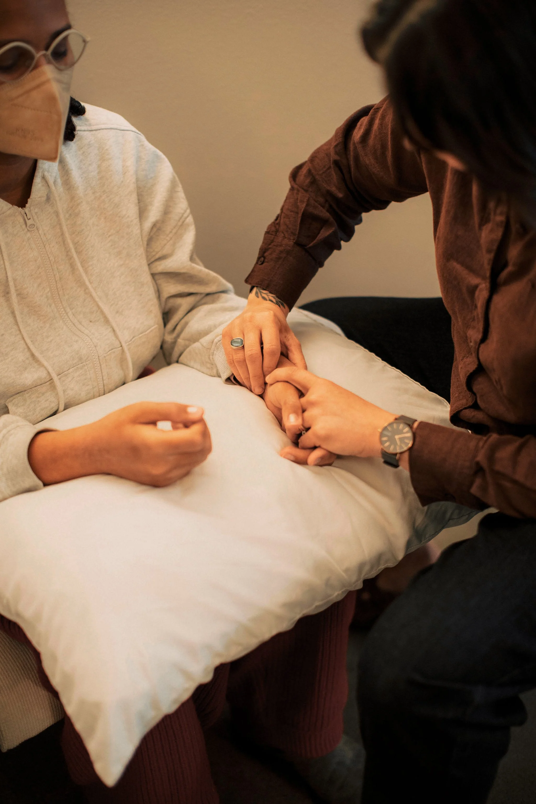 Two people, one wearing glasses and a beige sweater, and the other wearing a brown shirt, are holding hands on a white pillow. The person in the brown shirt has a tattoo on their wrist and is wearing a watch. The image appears to depict a moment of comforting or support.