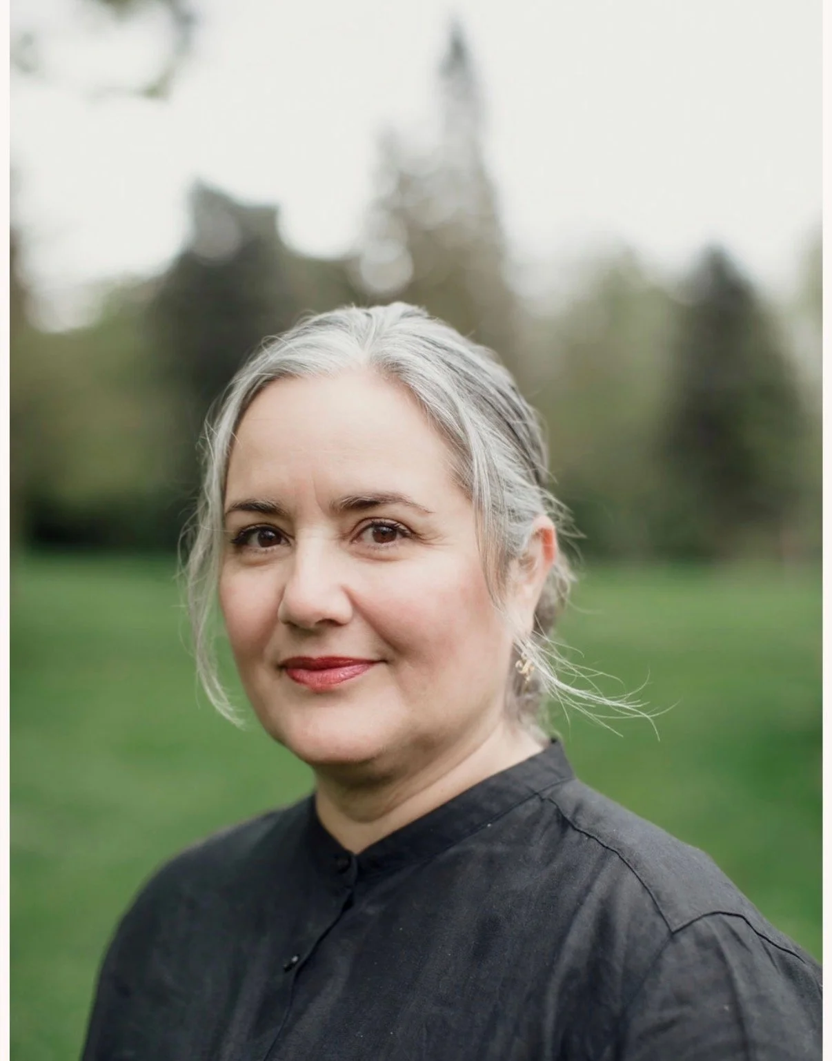 A portrait of a middle-aged woman with gray hair, wearing a black shirt, standing outdoors in a park with blurred trees in the background.