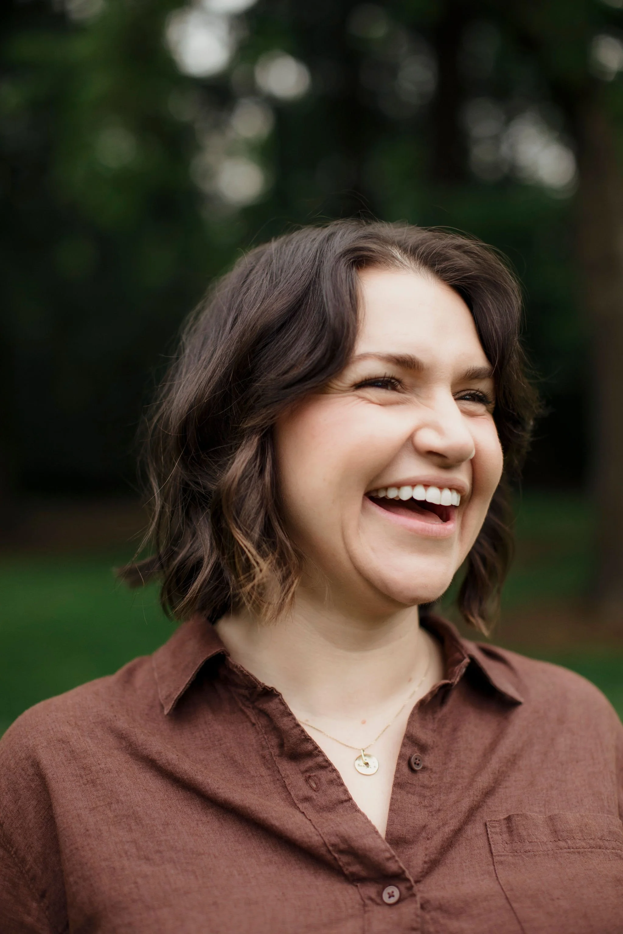 A woman with short, wavy brown hair, smiling outdoors with a background of blurred trees and greenery, wearing a brown button-up shirt and a gold necklace.