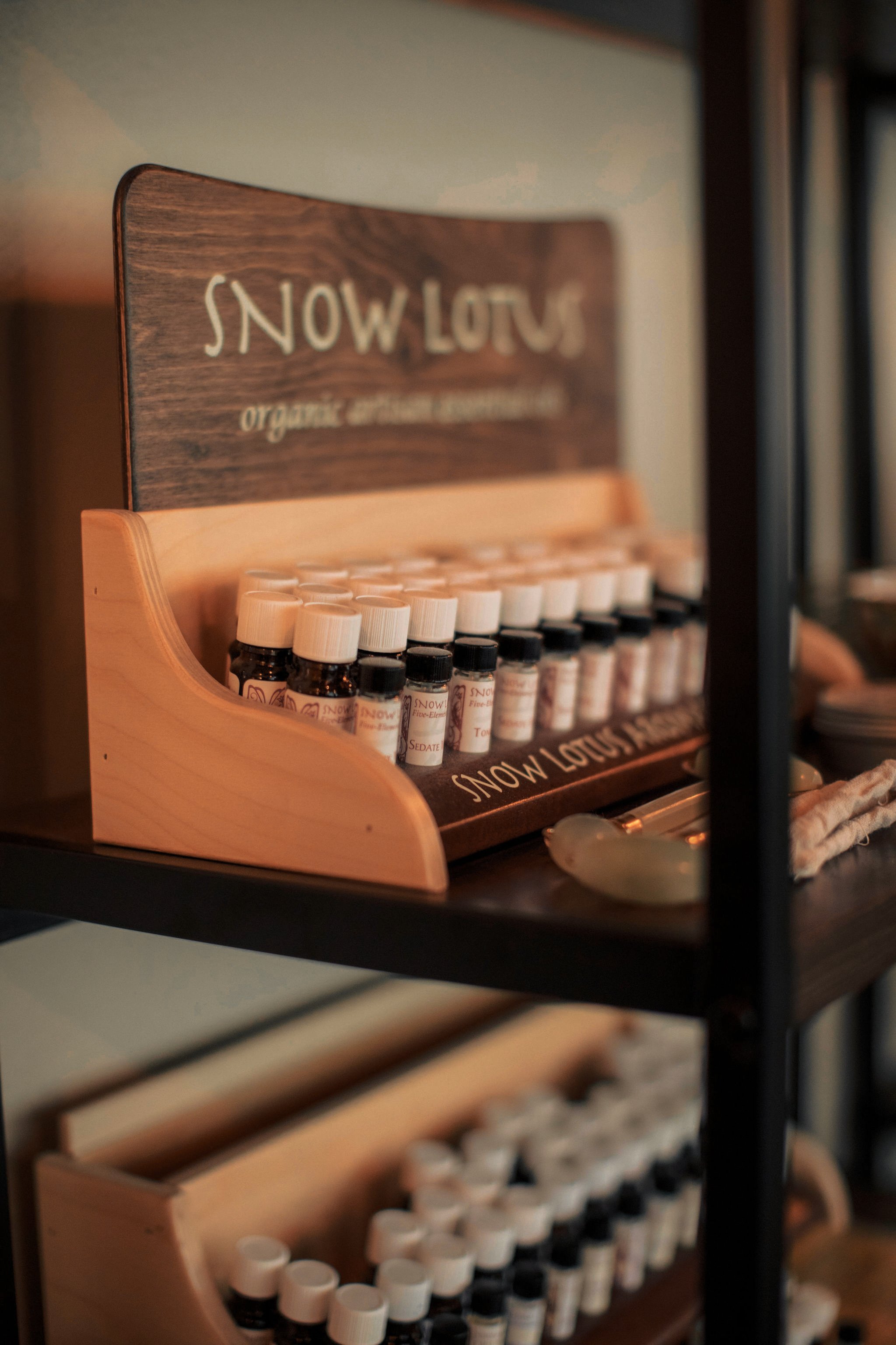 A wooden display stand with essential oils labeled Snow Lotus and various other labels, on a black metal shelf.