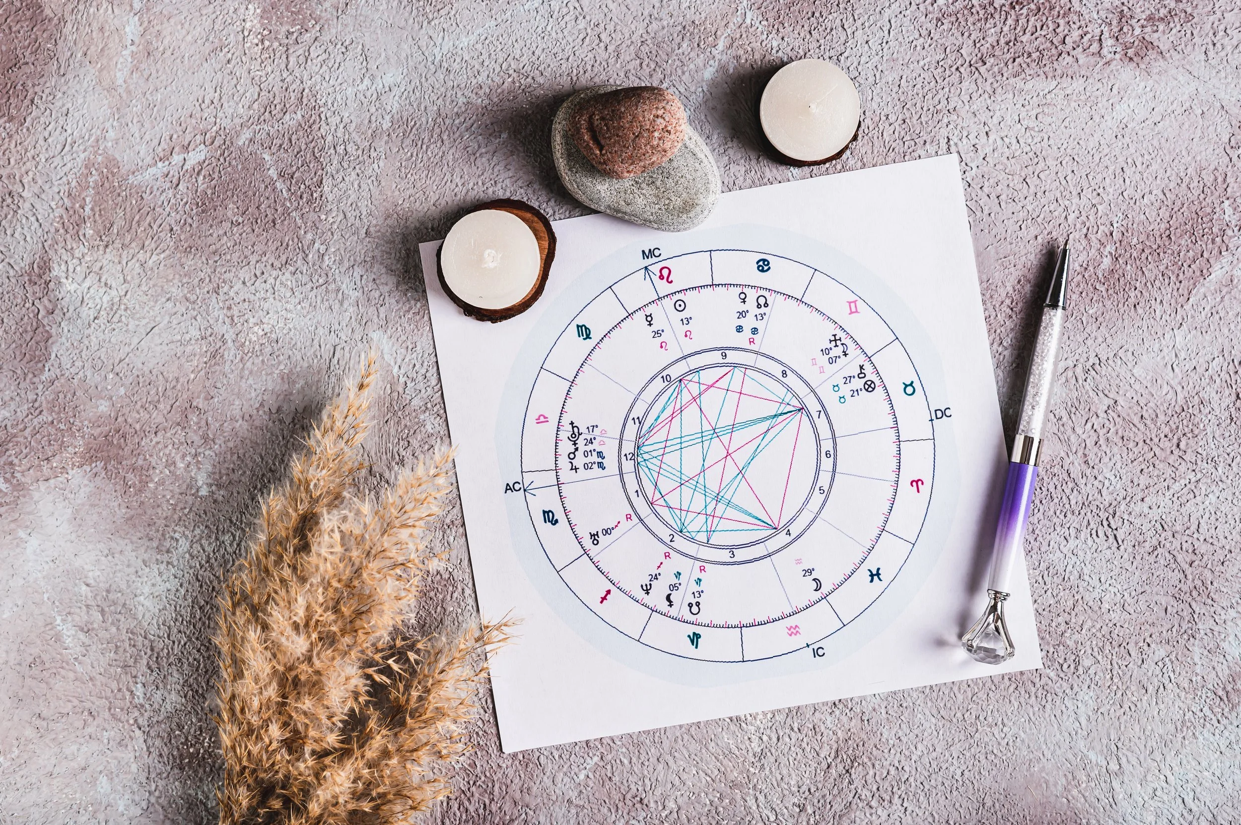 An astrology chart on a textured pink surface with three candles and three small rocks on top, alongside a silver pen with purple grip.