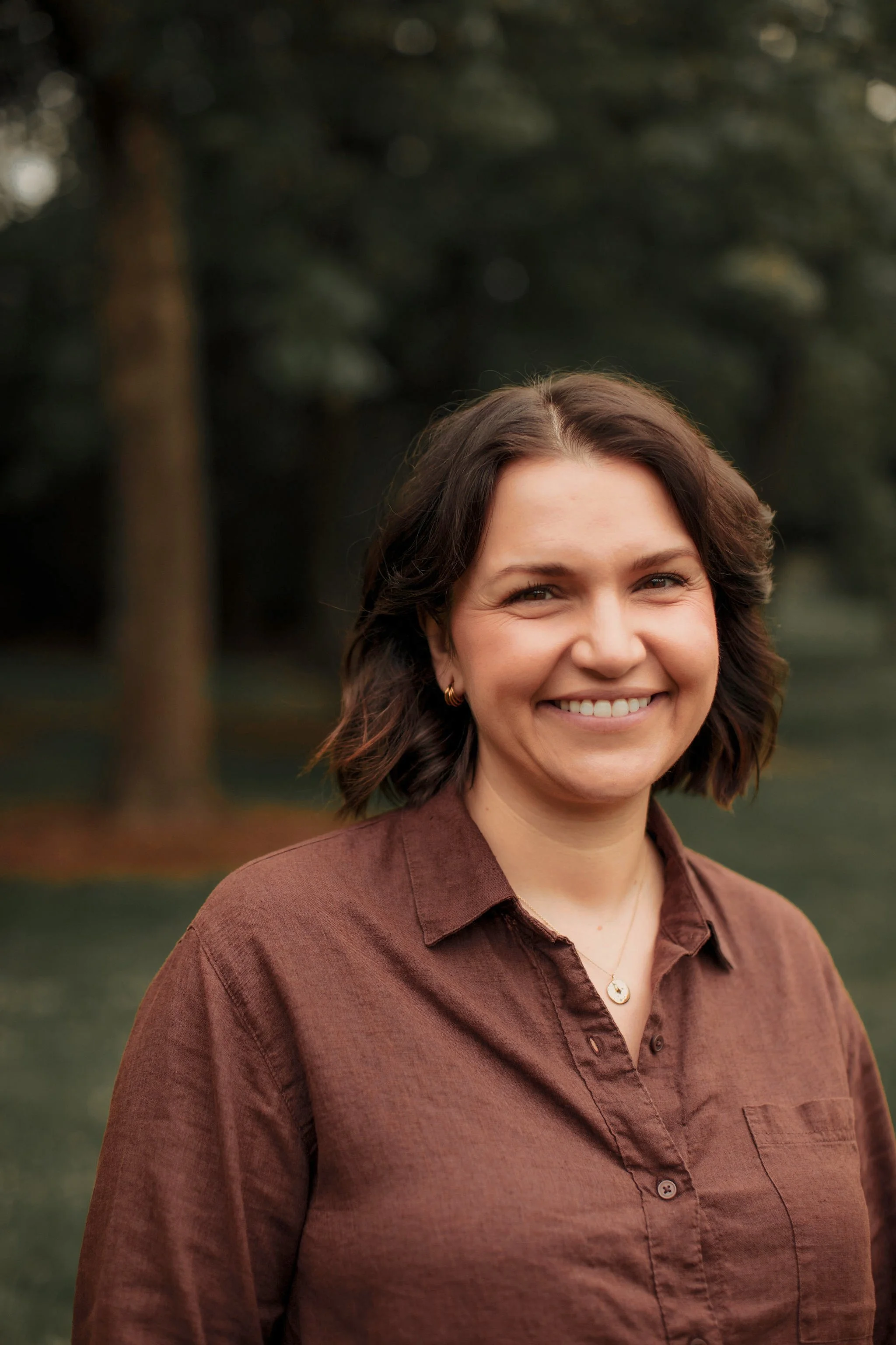 A woman with brown hair styled in loose waves, wearing a brown button-up shirt, smiling, outdoors with blurred trees in the background.
