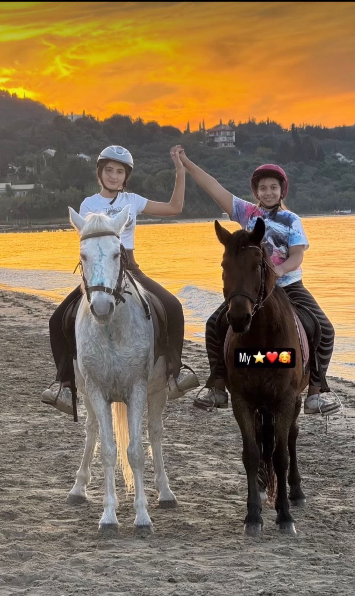 Two girls riding horses on a sandy beach during sunset, holding hands elevated in the air, with a scenic background of hills and houses, and a colorful sky.