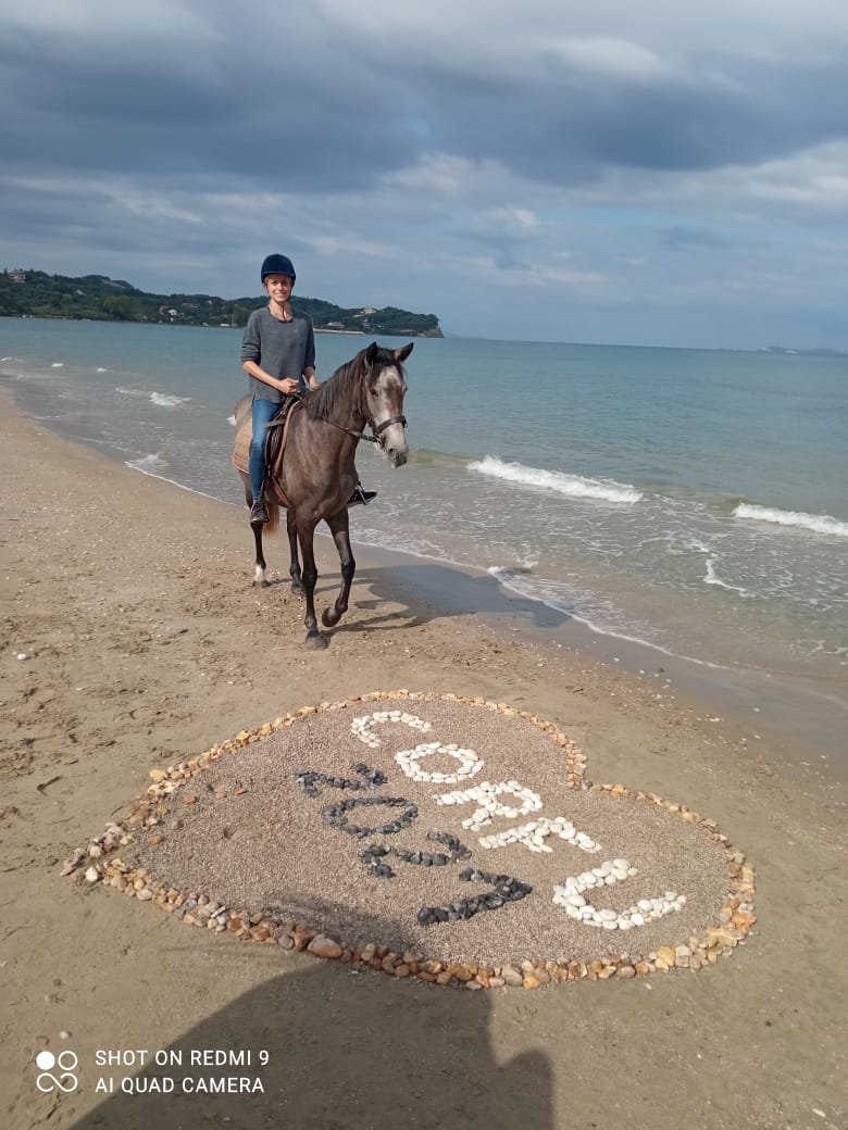 A woman riding a horse on a beach near the water, with a heart-shaped design made of rocks in the sand that reads "hope" and "Peace".