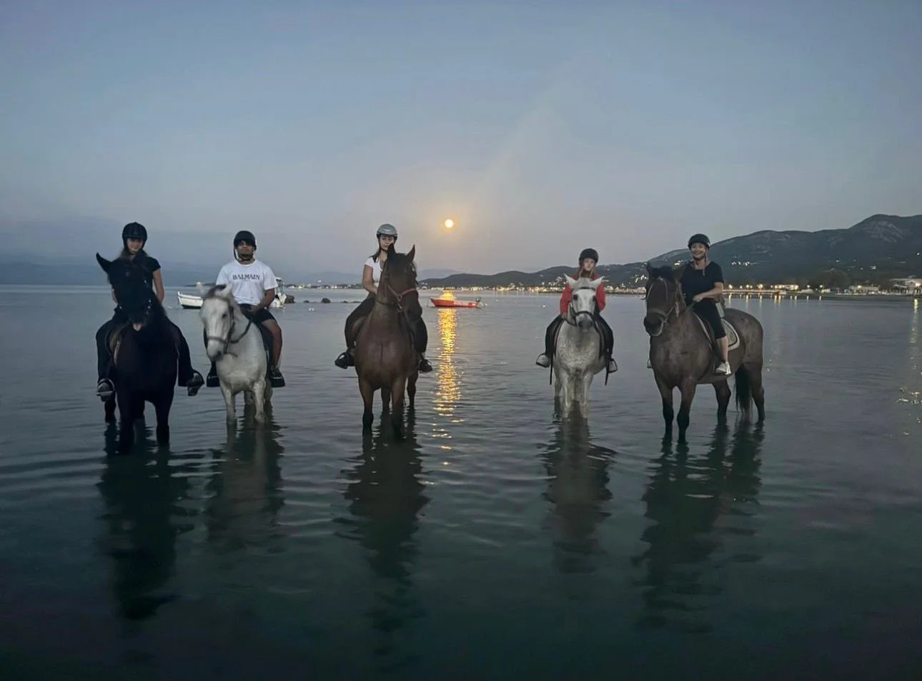 Five people riding horses in shallow water at sunset, with hills and boats in the background.