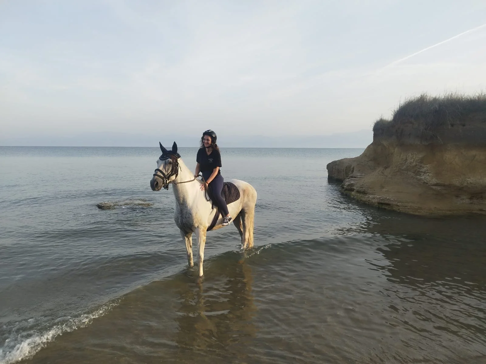 A woman riding a white horse in shallow water near a rocky shoreline and a cliff, with the ocean and sky in the background.