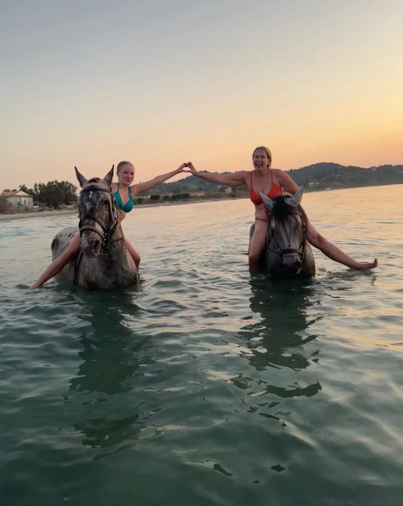 Two women riding horses in the water at sunset, holding hands to form a heart shape.