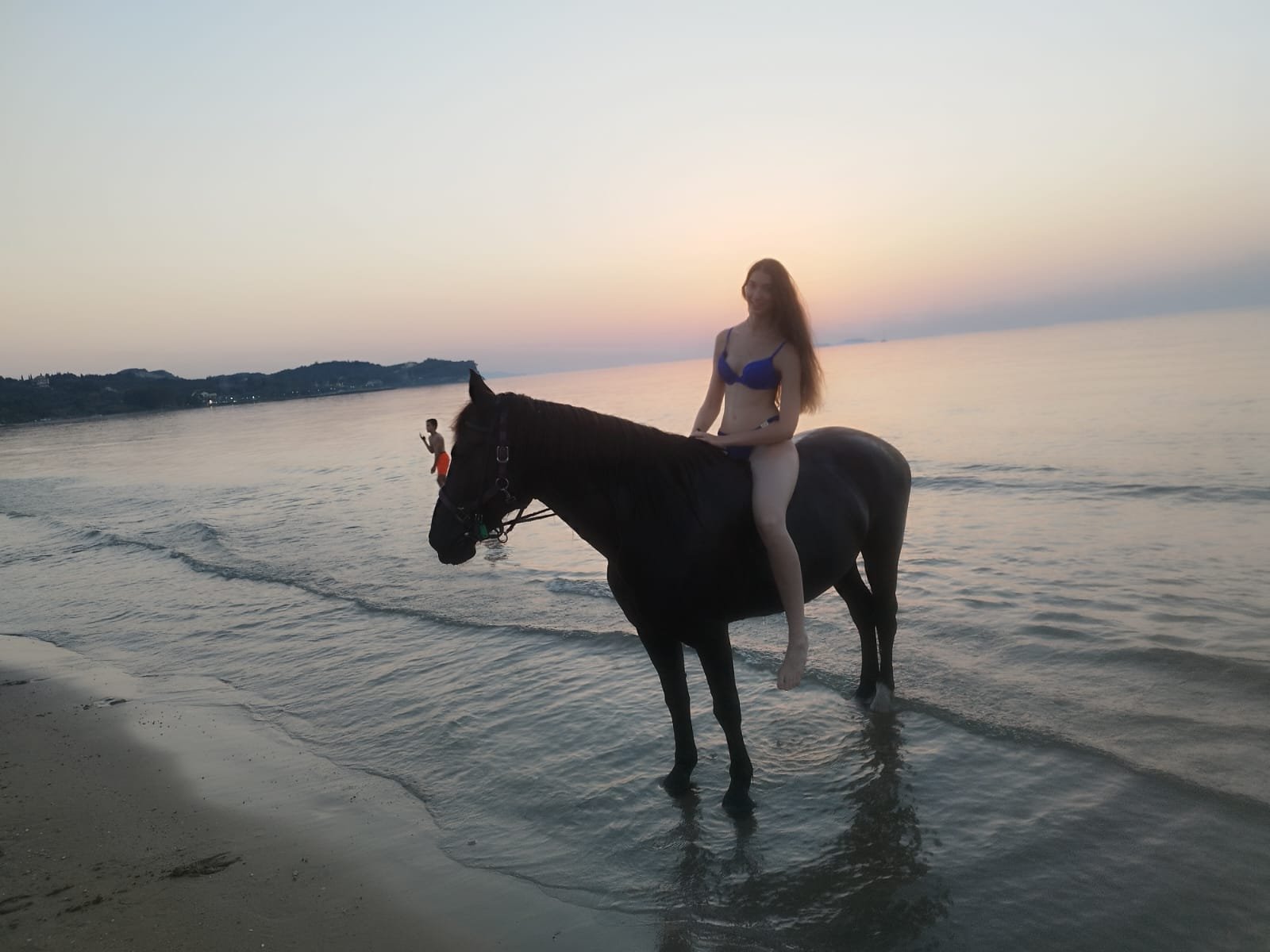 A woman in a blue bikini top and beige shorts riding a black horse in shallow water at the beach during sunset, with a child in the background near the shoreline.