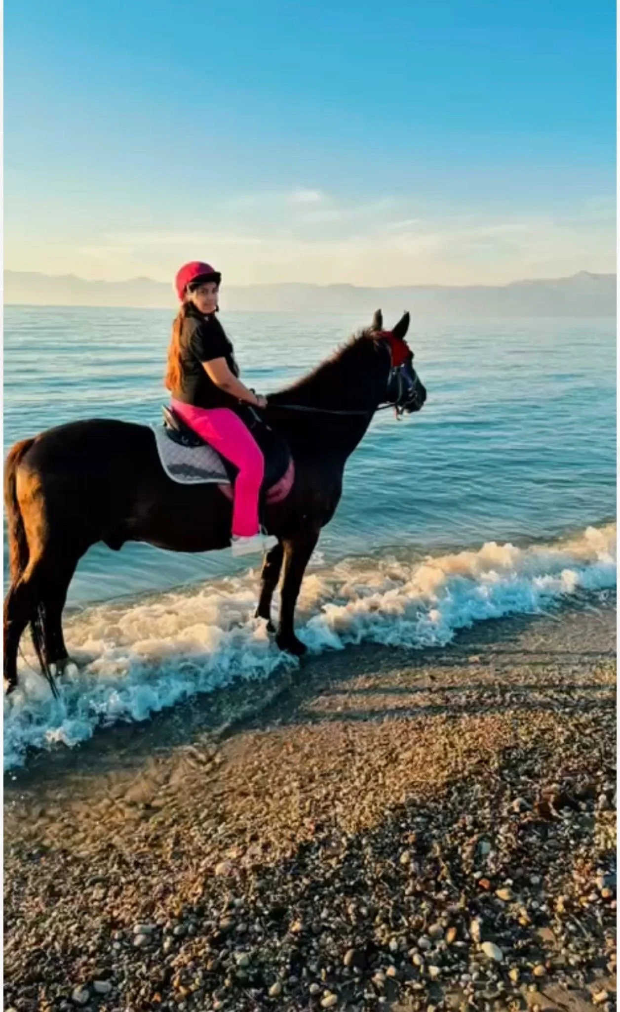 Woman riding a black horse along the shoreline with waves in the background and distant mountains visible on the horizon during sunset.