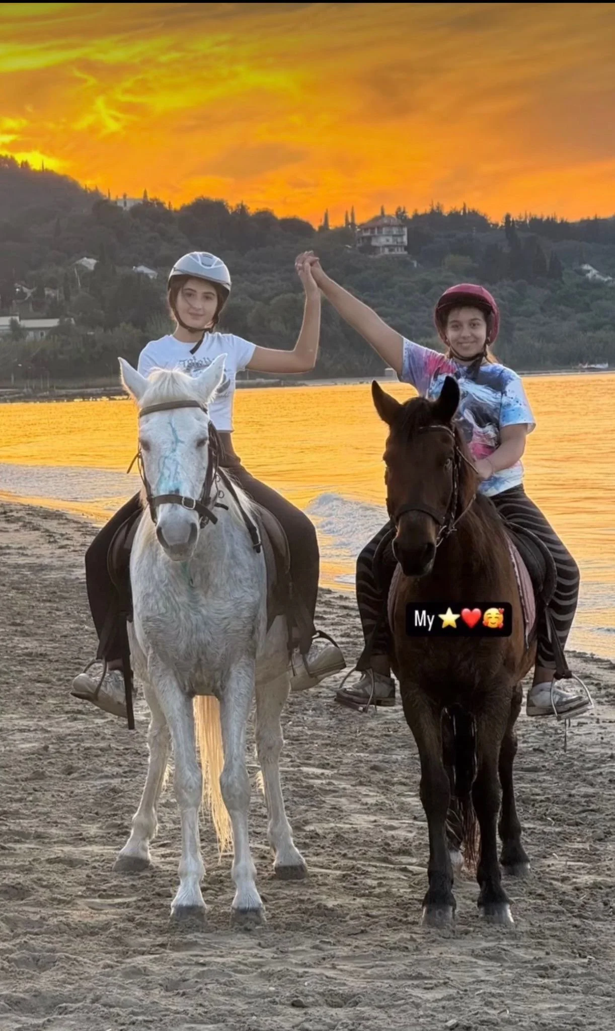 Two children riding horses along a beach at sunset, with one child wearing a white helmet and the other a red helmet, and holding hands in the air. The background features a sunset sky with orange and yellow clouds, hills with houses, and water refle