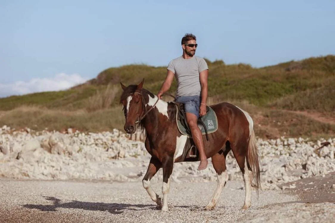 A man riding a brown and white horse on a beach with grassy hills in the background.