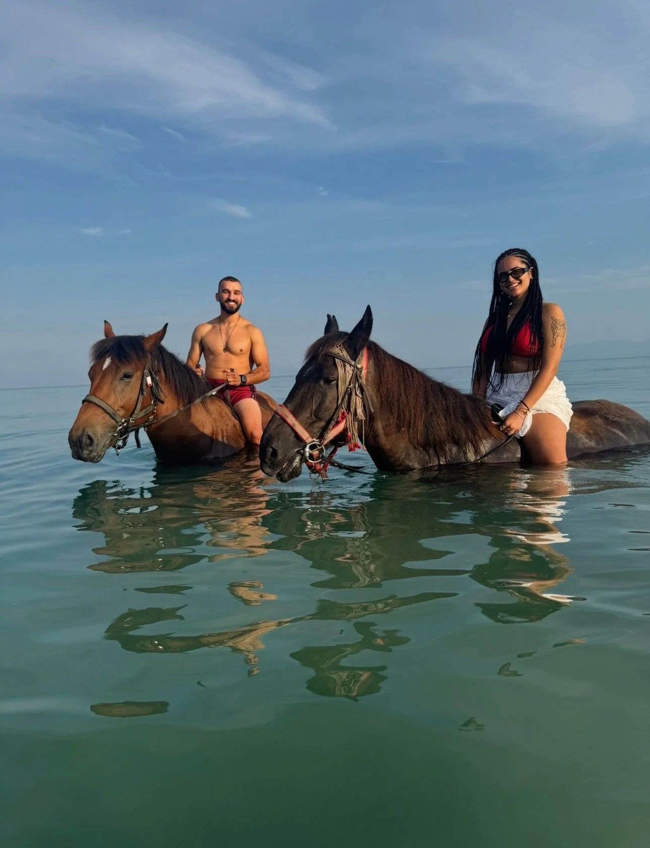 Two people riding horses in water, a man on the left and a woman on the right, under a blue sky with clouds.