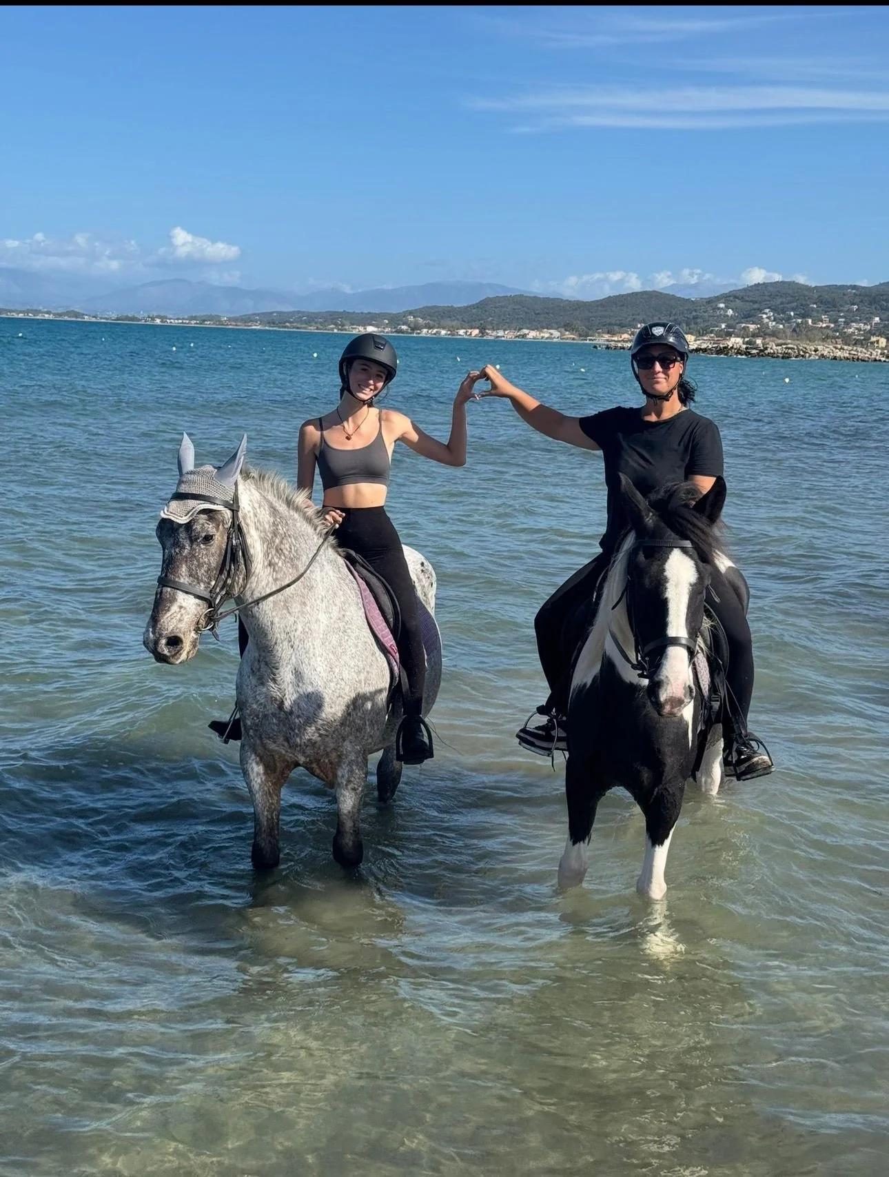 Two women riding horses in the ocean, holding hands with fingers touching, wearing helmets and black riding clothes. The horses are standing in shallow water by the beach, with a distant shoreline, mountains, and blue sky in the background.