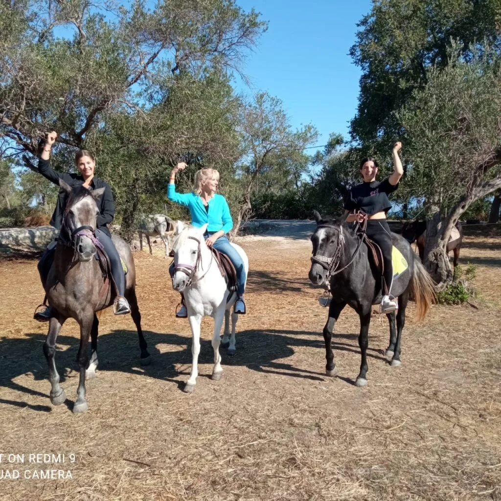 Three women riding horses outdoors on a sunny day, in a natural setting with trees and clear blue sky, with each woman flexing one arm in a celebratory gesture.