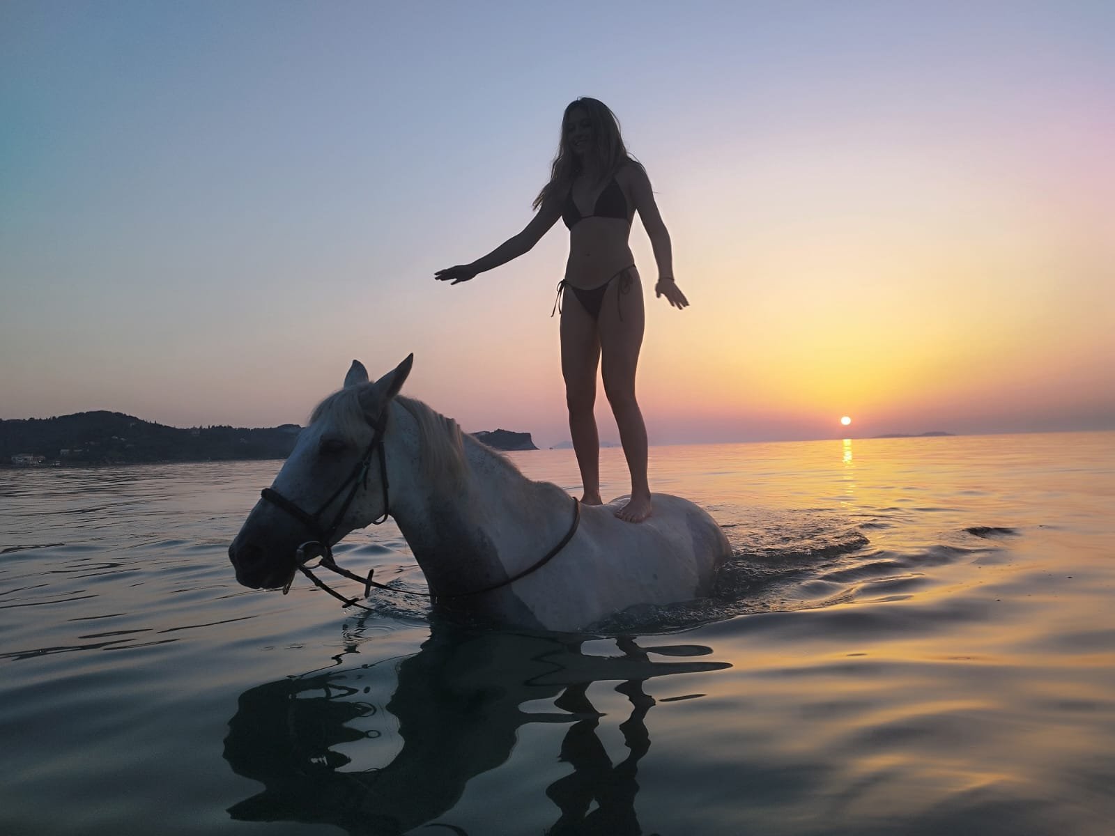 A person in a bikini standing on a white horse in the water during sunset.