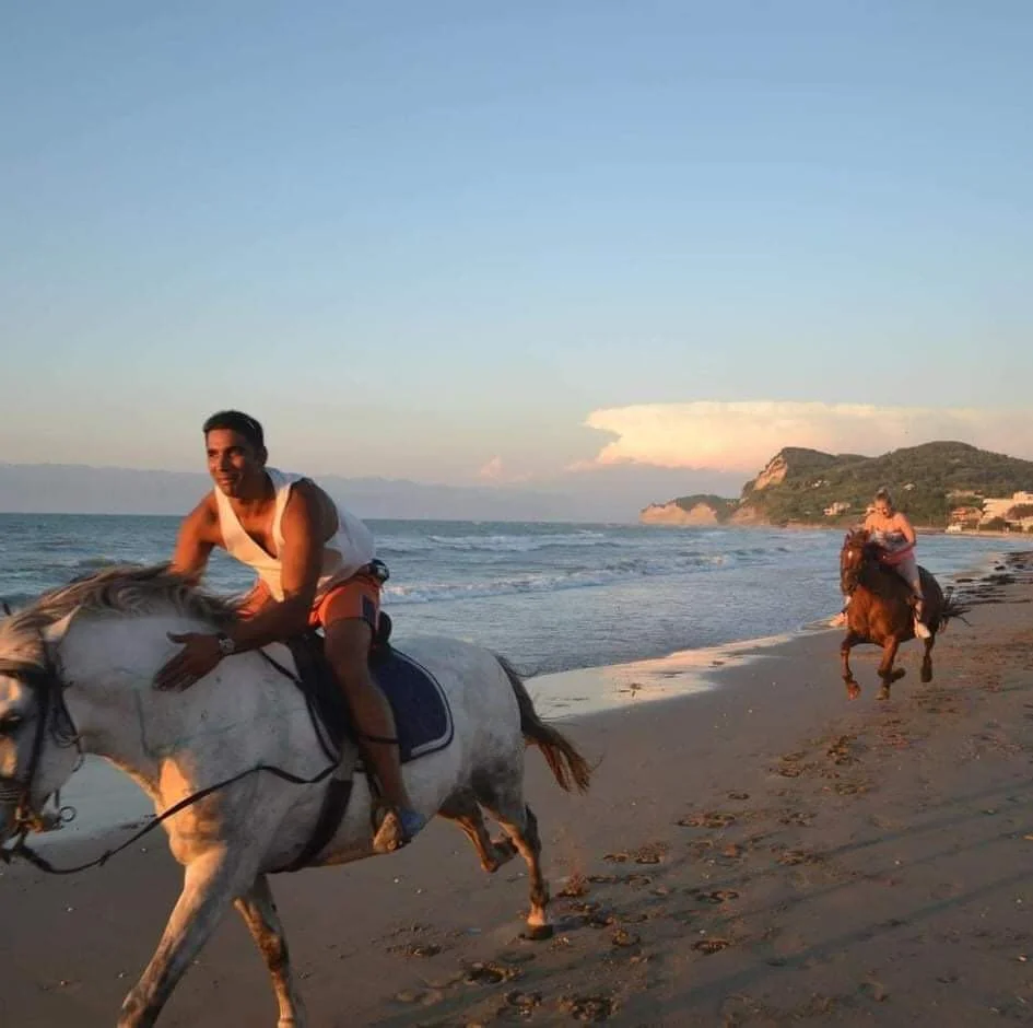Two people riding horses on a beach during sunset with ocean waves and a distant hillside.